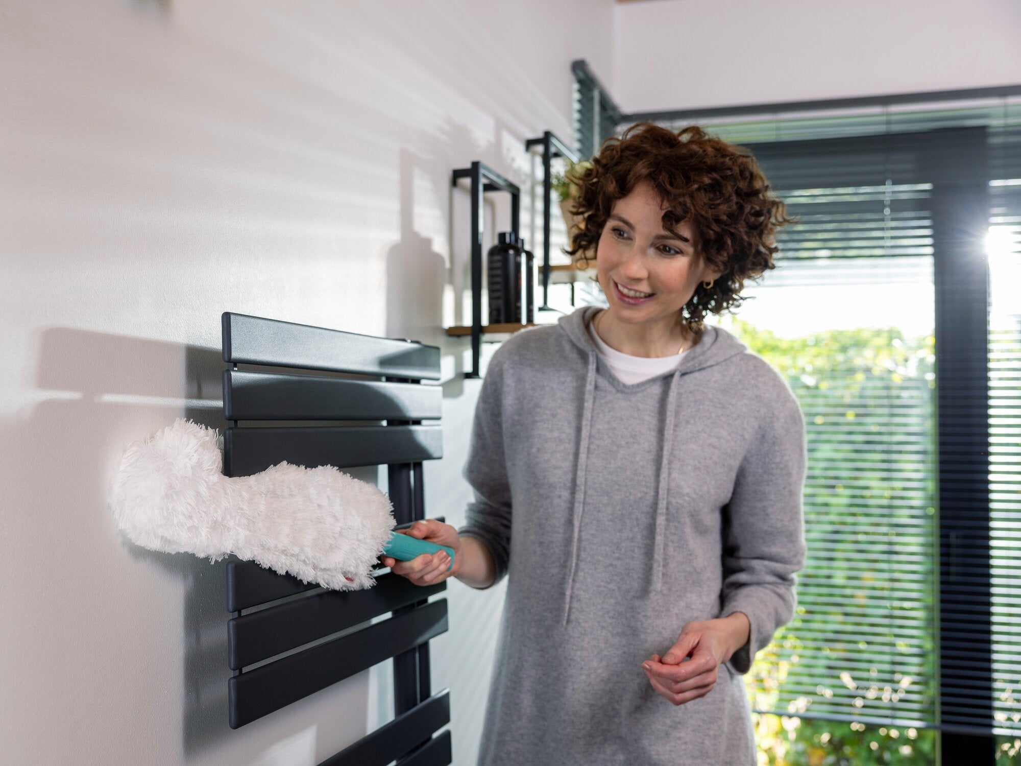 Una mujer utiliza el plumero SUPERDUSTER XL para limpiar un radiador en una habitación moderna. El plumero tiene una cabeza grande y esponjosa, ideal para lugares de difícil acceso.