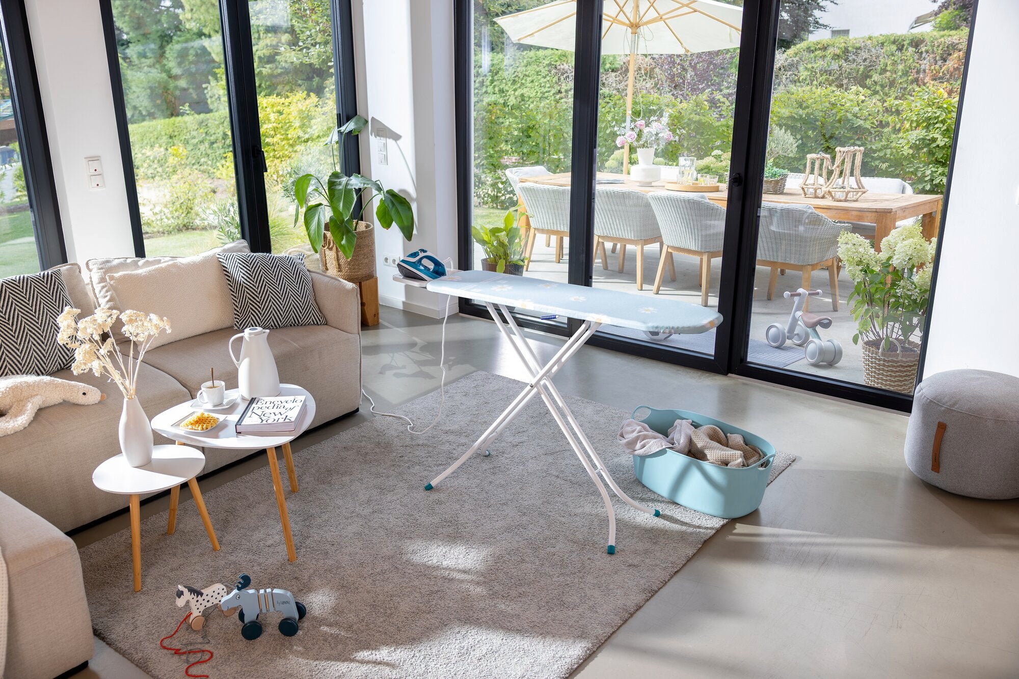 Bügelbrett Leifheit Classic S Basic in einem modernen Wohnzimmer mit großen Fenstern. Das Brett steht auf einem Teppich, umgeben von Spielzeug und Pflanzen, mit Blick auf einen sonnigen Garten.