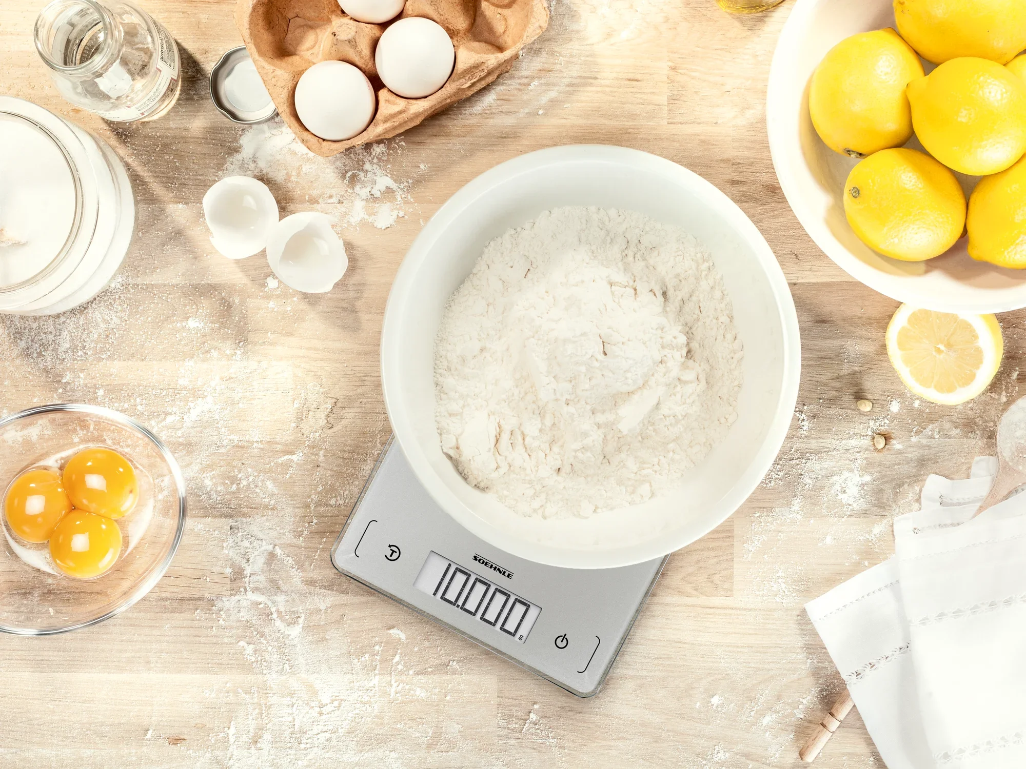 Digitale keukenweegschaal Page Comfort 300 Slim op een werkblad met bloem, eieren en citroenen. De weegschaal toont het gewicht van de ingrediënten in een kom. Ideaal voor nauwkeurig bakken.