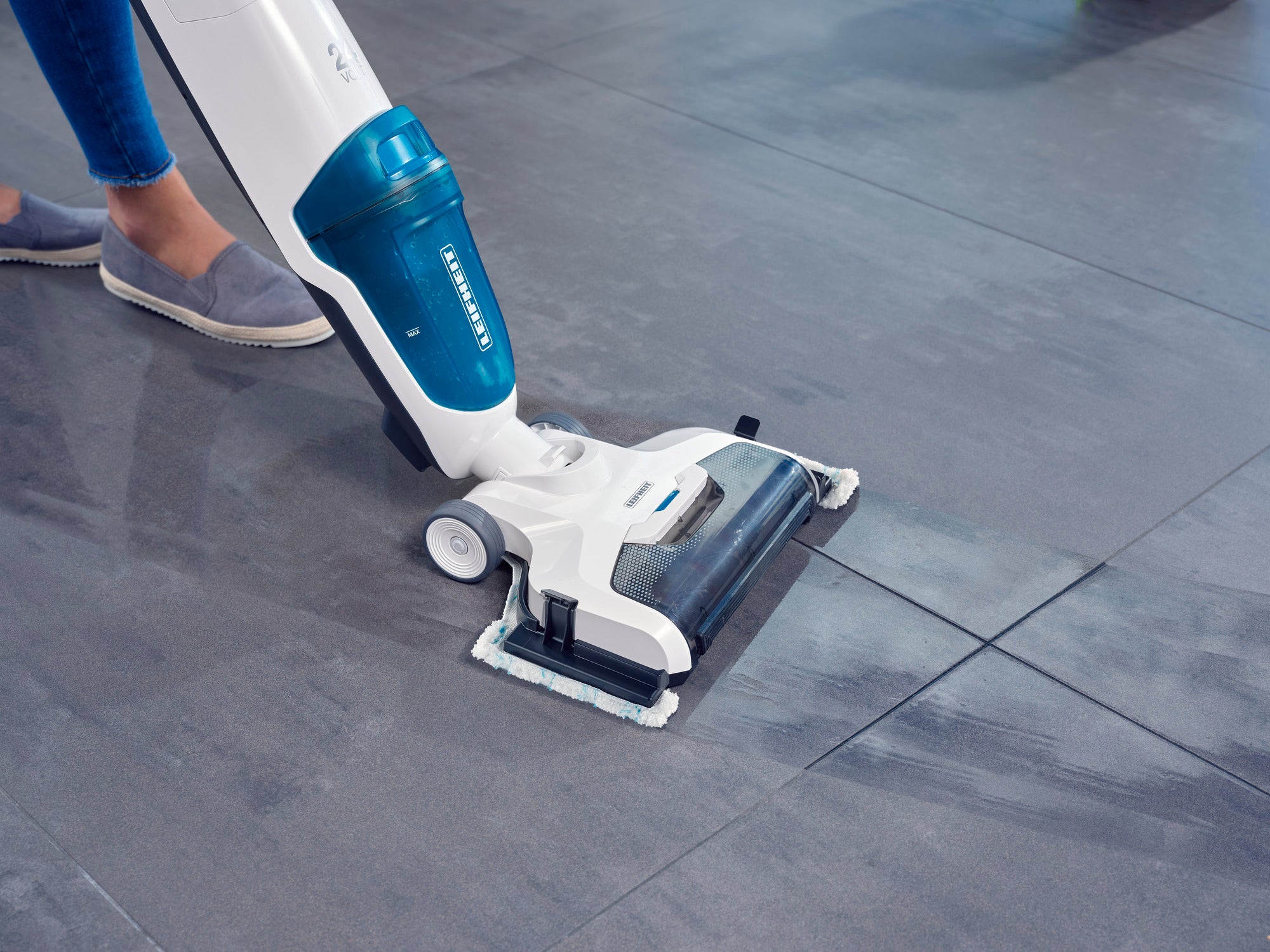 Aspirateur Regulus Aqua PowerVac Pro en action sur un sol carrelé gris, montrant de près sa capacité à nettoyer efficacement. Une personne en baskets l'utilise.