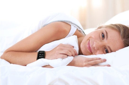 Eine laechelnde Frau liegt entspannt im Bett, eingehuellt in weisse Bettwaesche, waehrend sie eine Smartwatch am Handgelenk traegt.