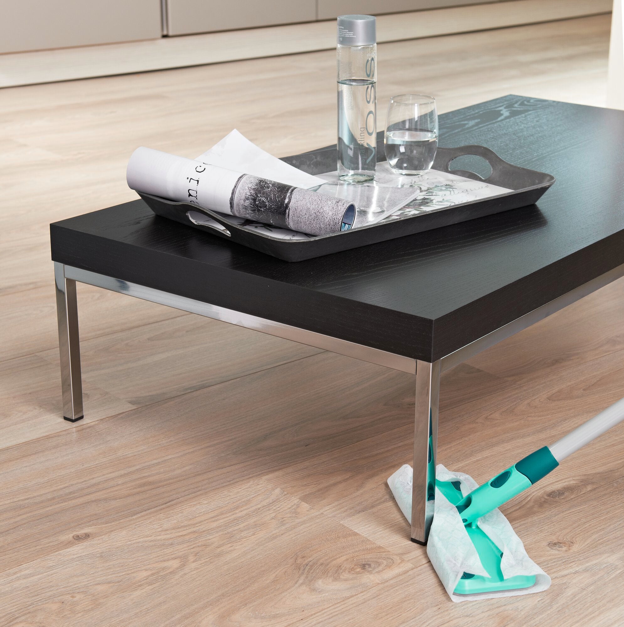 Bodenwischer Clean&Away in einem modernen Wohnzimmer. Der Wischer liegt auf einem hellen Holzboden neben einem schwarzen Couchtisch mit Wasserflasche und Zeitung.