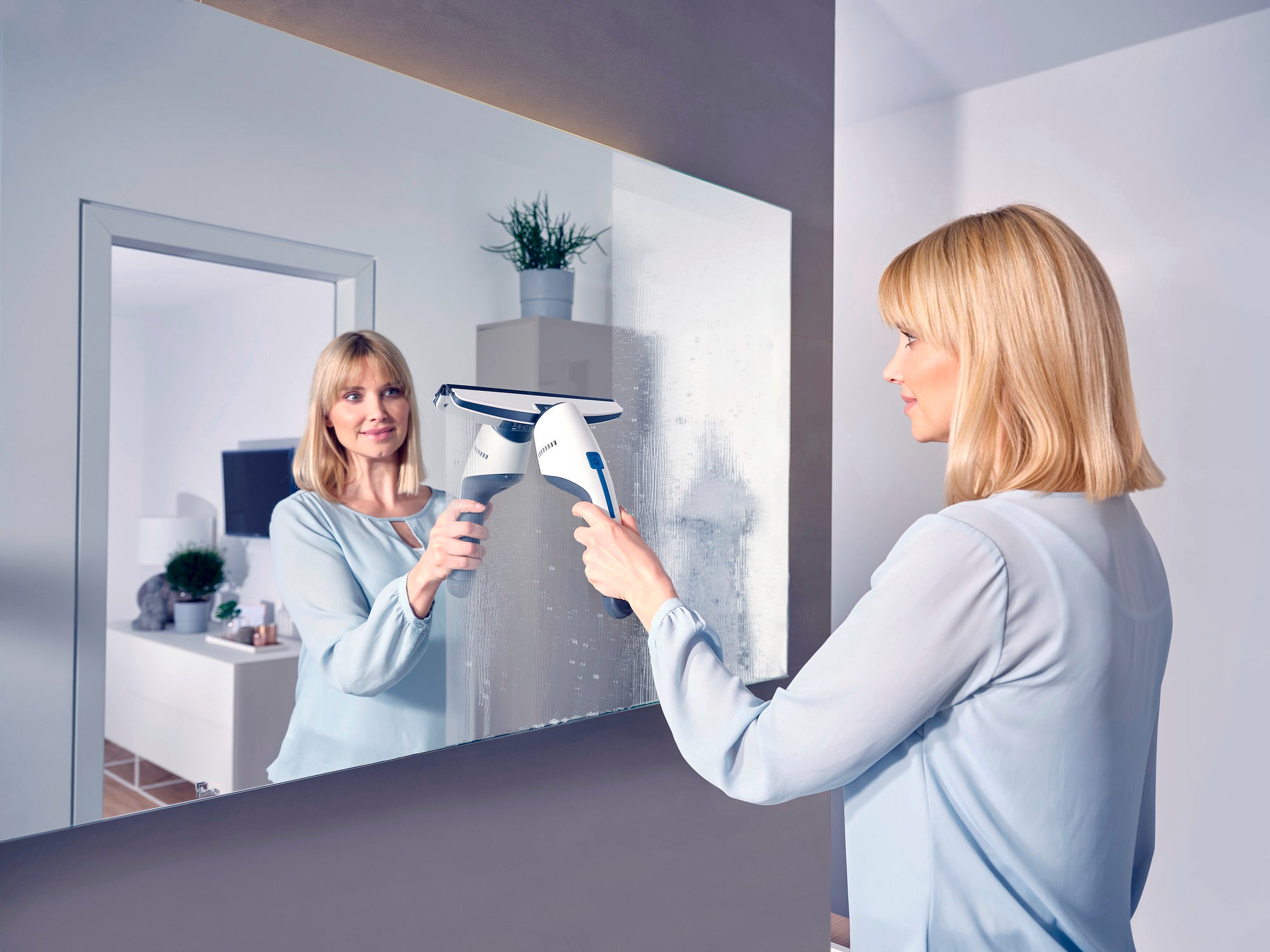 Een vrouw maakt een spiegel schoon met de Nemo raamstofzuiger. De stofzuiger verwijdert effectief water en vuil terwijl de vrouw naar haar spiegelbeeld kijkt. Ideaal voor streeploos schoon.