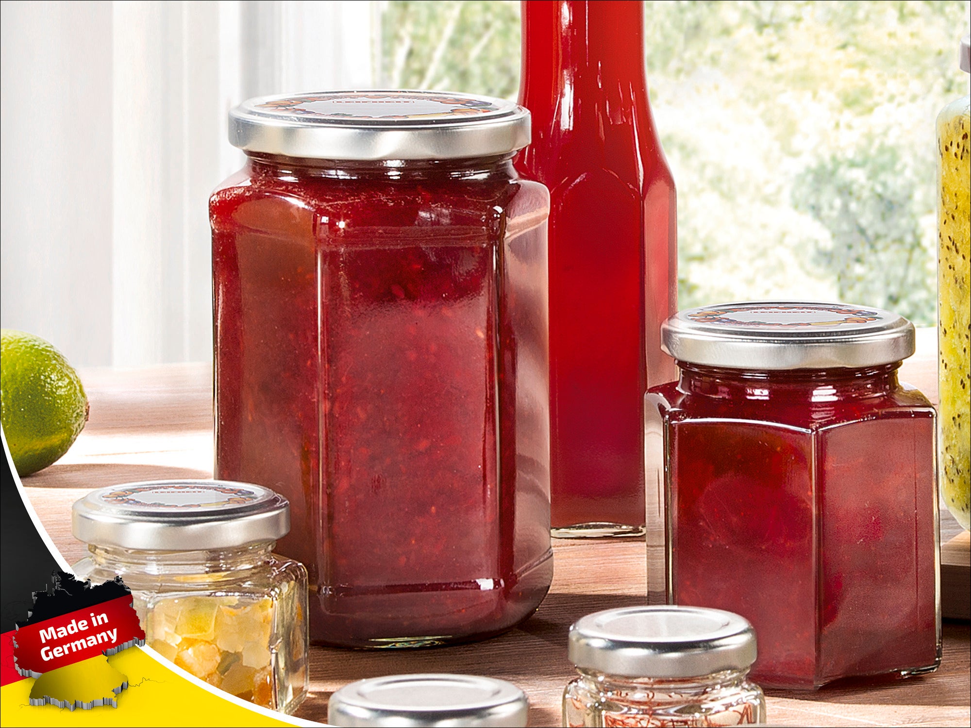 Un pot contenant de la confiture rouge est posé sur une table, entouré d'autres pots contenant différents produits. À l'arrière-plan, on aperçoit une bouteille de jus rouge. Une étiquette « Made in Germany » est visible.