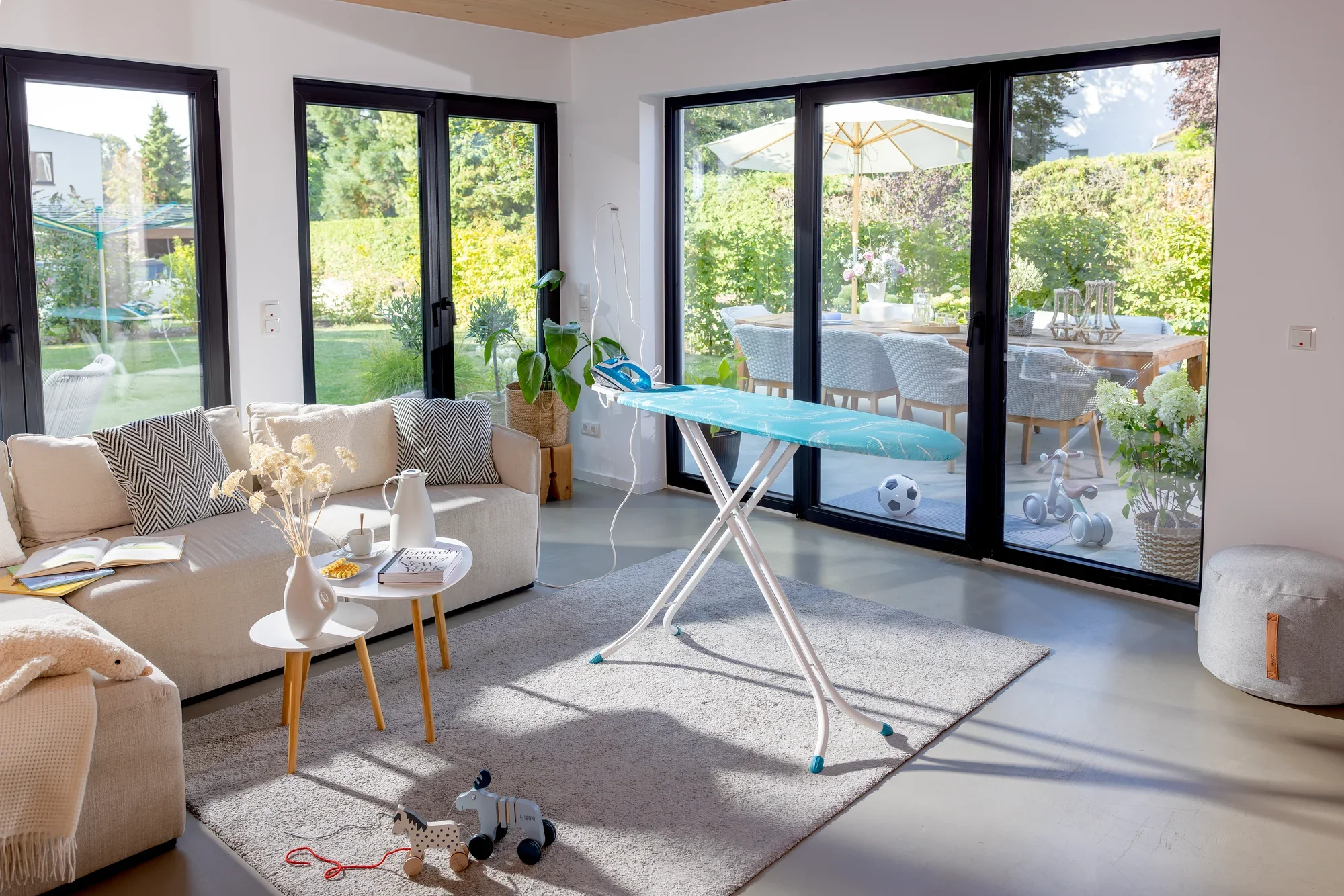 Bügelbrett Leifheit Air Board M Compact Plus in einem modernen Wohnzimmer mit großen Fenstern. Das Brett steht auf einem Teppich, umgeben von einem Sofa und einem Tisch.