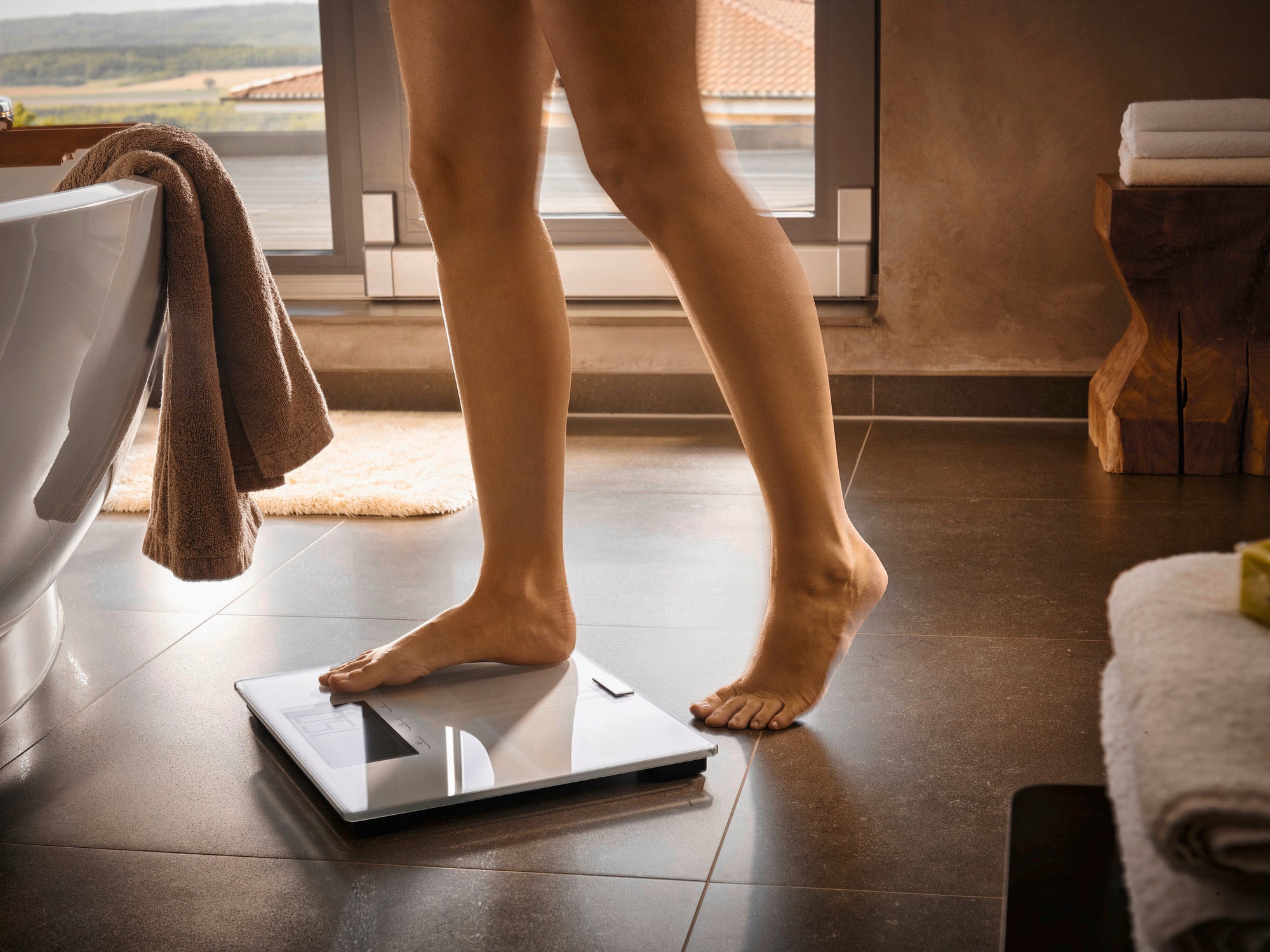 Osoba stoi na cyfrowej wadze łazienkowej Shape Sense Connect 200 w nowoczesnej łazience. Waga ma elegancki wygląd i odbija podłogę.