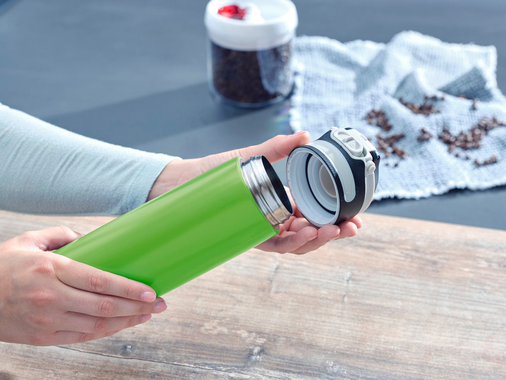 Isolierbecher Flip 600ml in Kiwi-Grün wird geöffnet. Der Becher liegt auf einem Holztisch, im Hintergrund sind Kaffeebohnen und ein weißes Tuch zu sehen. Ideal für unterwegs.