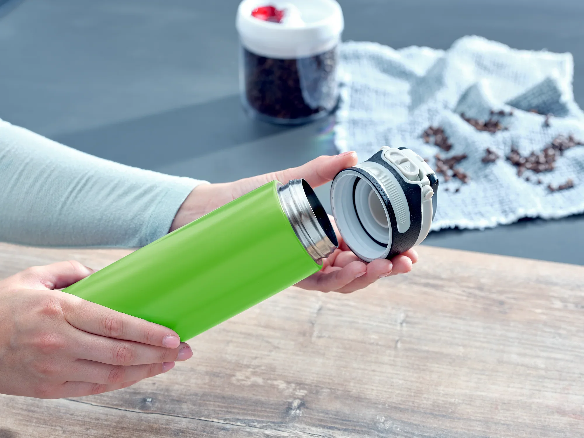 Kubek próżniowy Flip 600 ml w kolorze kiwi jest otwarty. Kubek leży na drewnianym stole, a w tle widać ziarna kawy i biały materiał. Idealny w podróży.