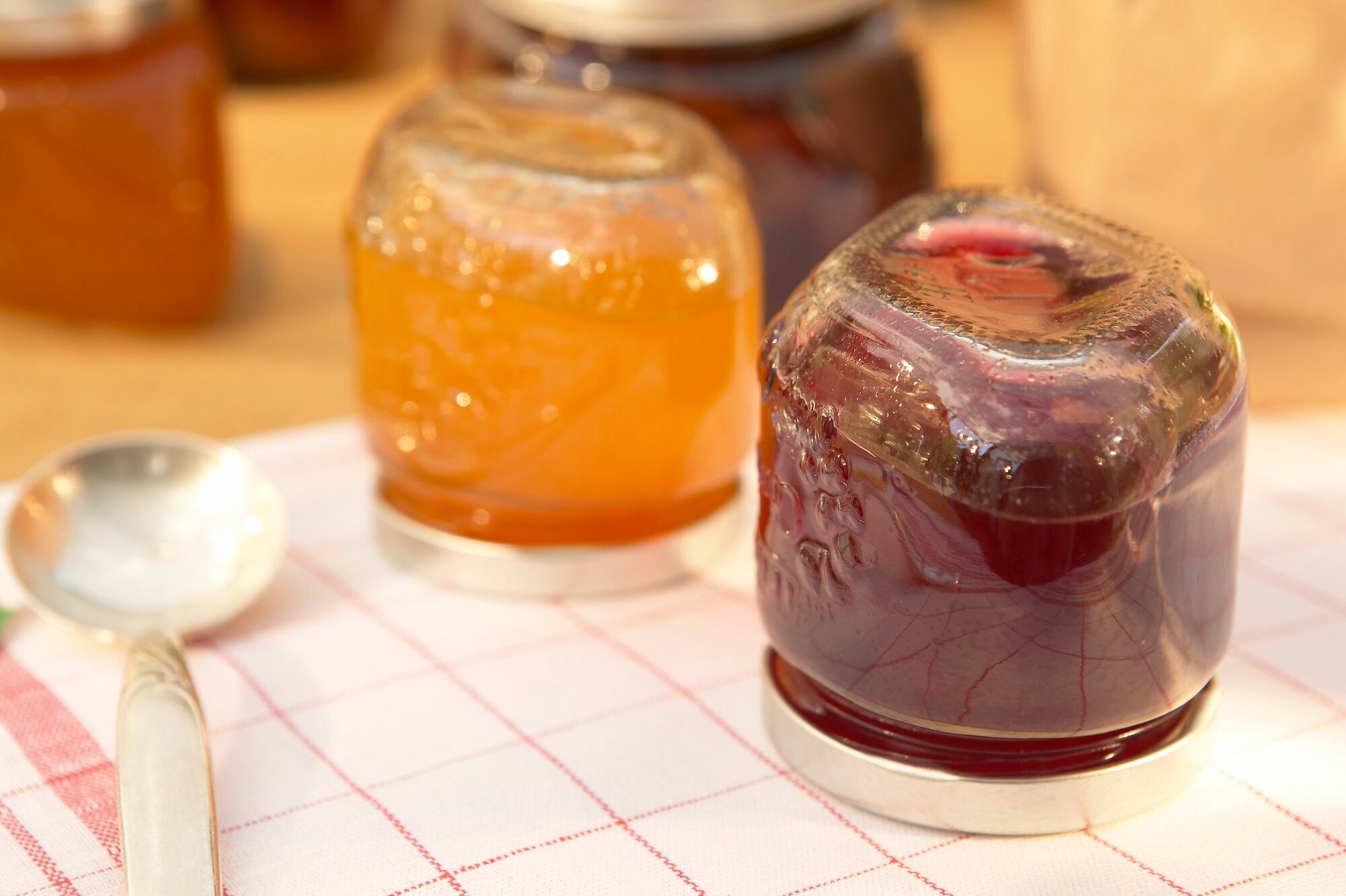 Zwei Gläser Gelee, eines mit orangefarbenem und eines mit dunkelrotem Inhalt, stehen auf einem karierten Tuch. Im Hintergrund sind weitere Gläser zu sehen.