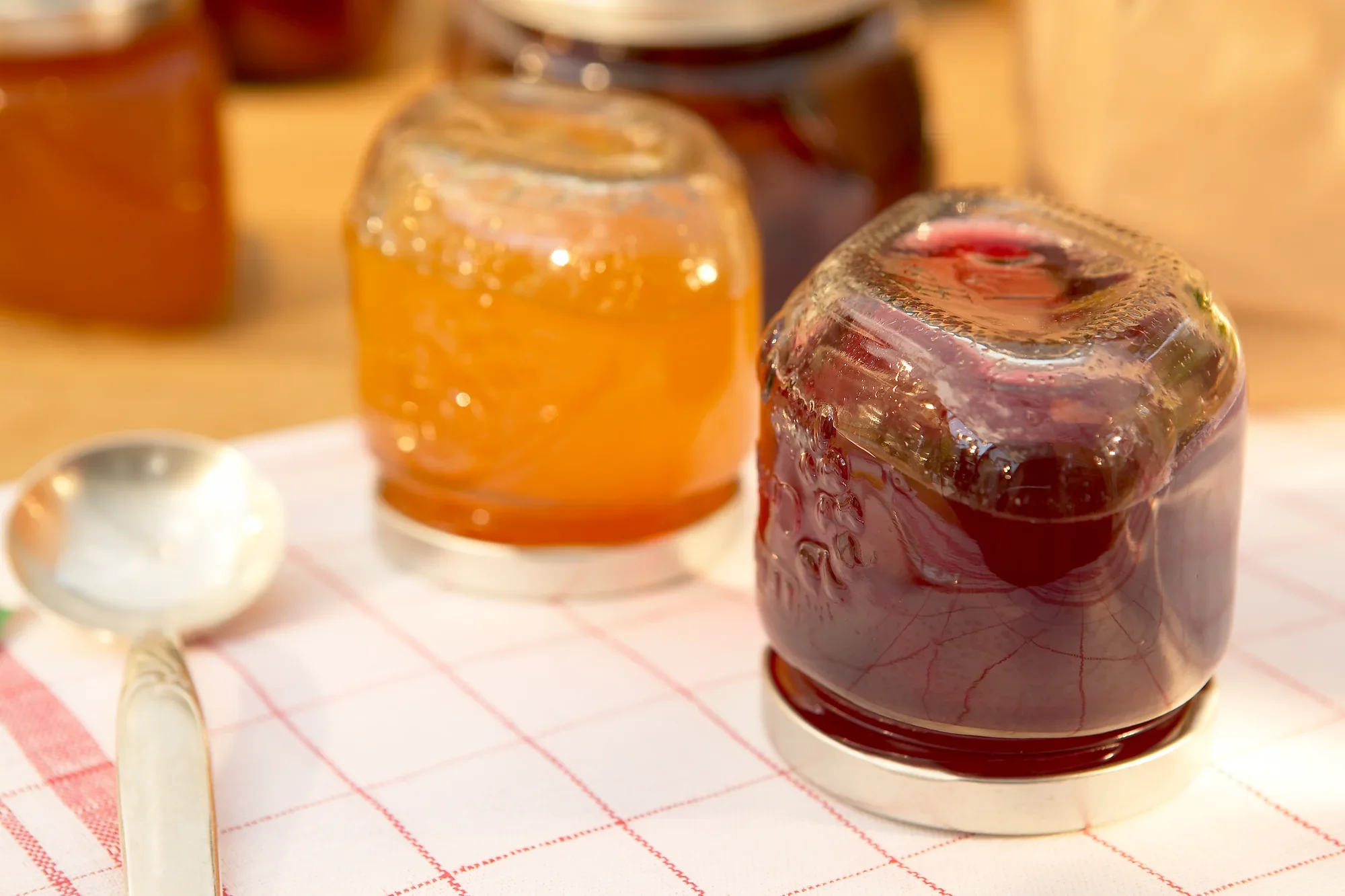 Dwa słoiki galaretki, jeden z pomarańczową, a drugi z ciemnoczerwoną zawartością, stoją na kraciastym obrusie. W tle widać więcej słoików.