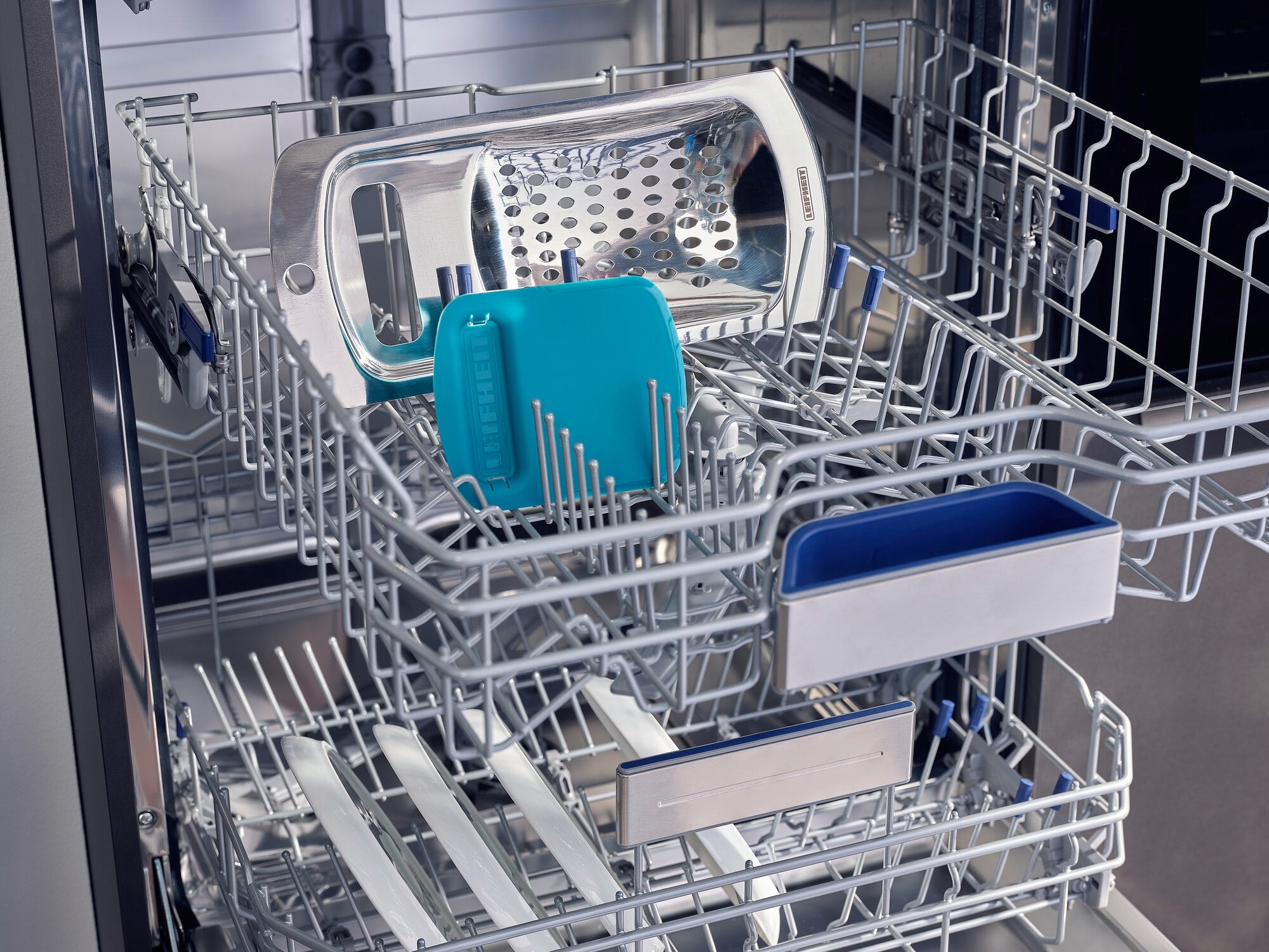 Spaetzle schraper en rasp in één vaatwasser. De spätzle schraper is turquoise en zit naast een zilveren rasp op het bovenste schap van de vaatwasser.