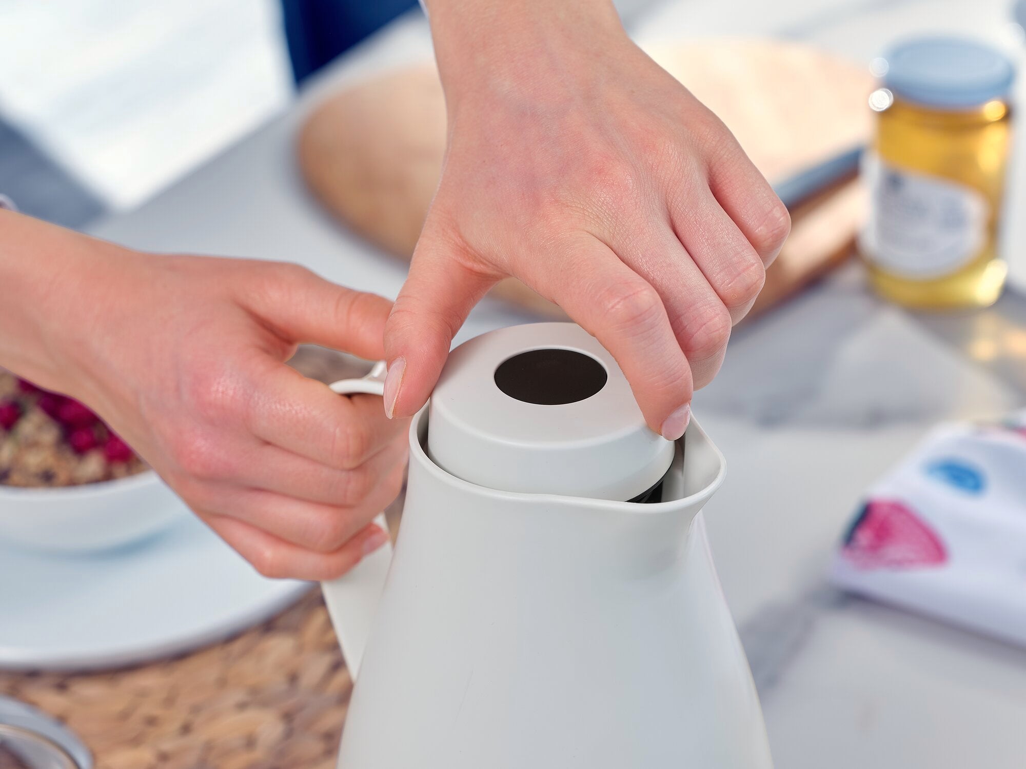 Een persoon opent het deksel van de Harmonic isoleerkan in een keuken. Op de achtergrond zijn een snijplank, een kom muesli en een pot honing te zien.