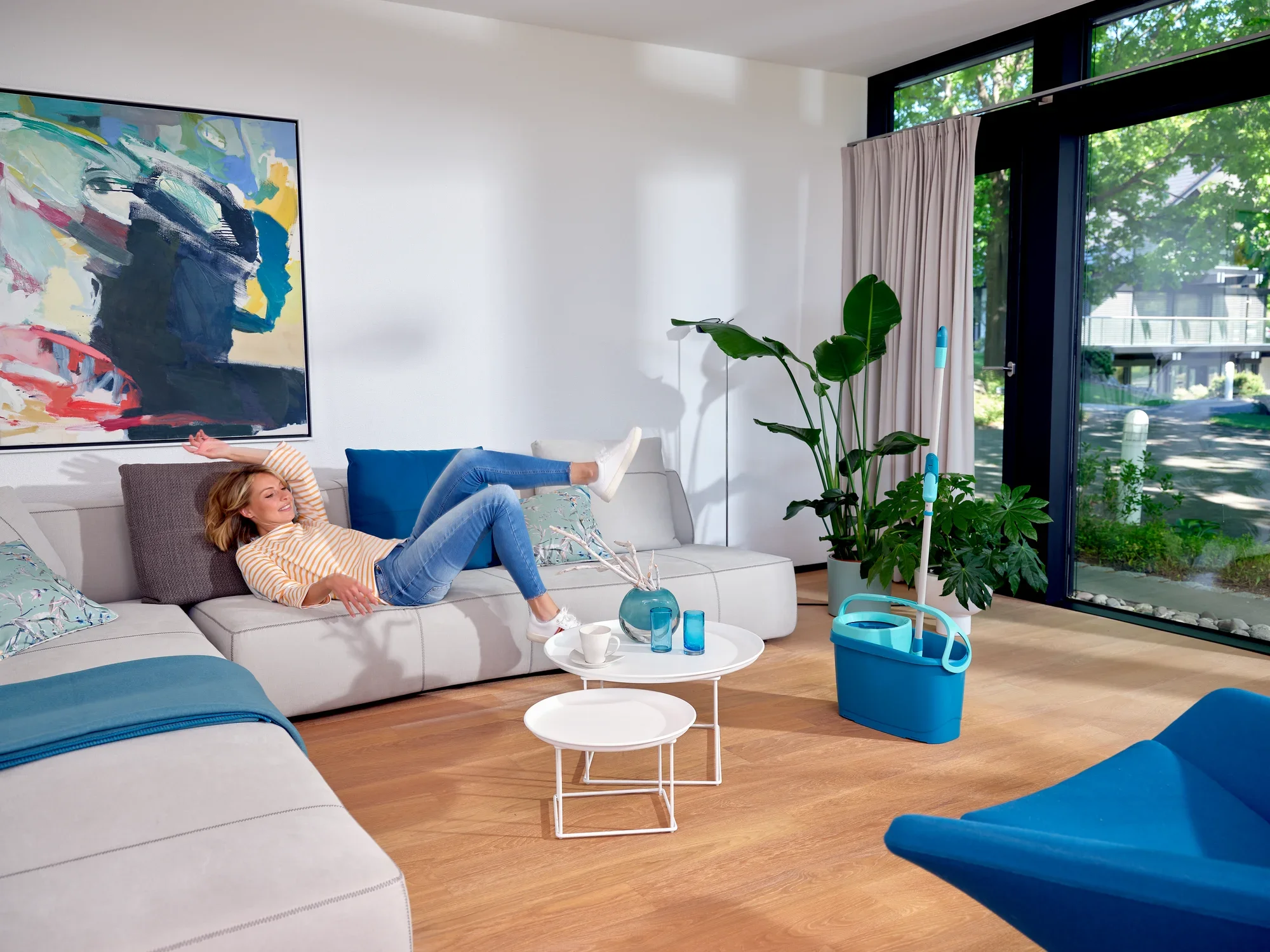 Eine Frau entspannt sich auf einem modernen Sofa in einem hellen Wohnzimmer. Neben ihr steht das CLEAN TWIST Set mit Eimer und Wischmopp, bereit für die nächste Reinigung.