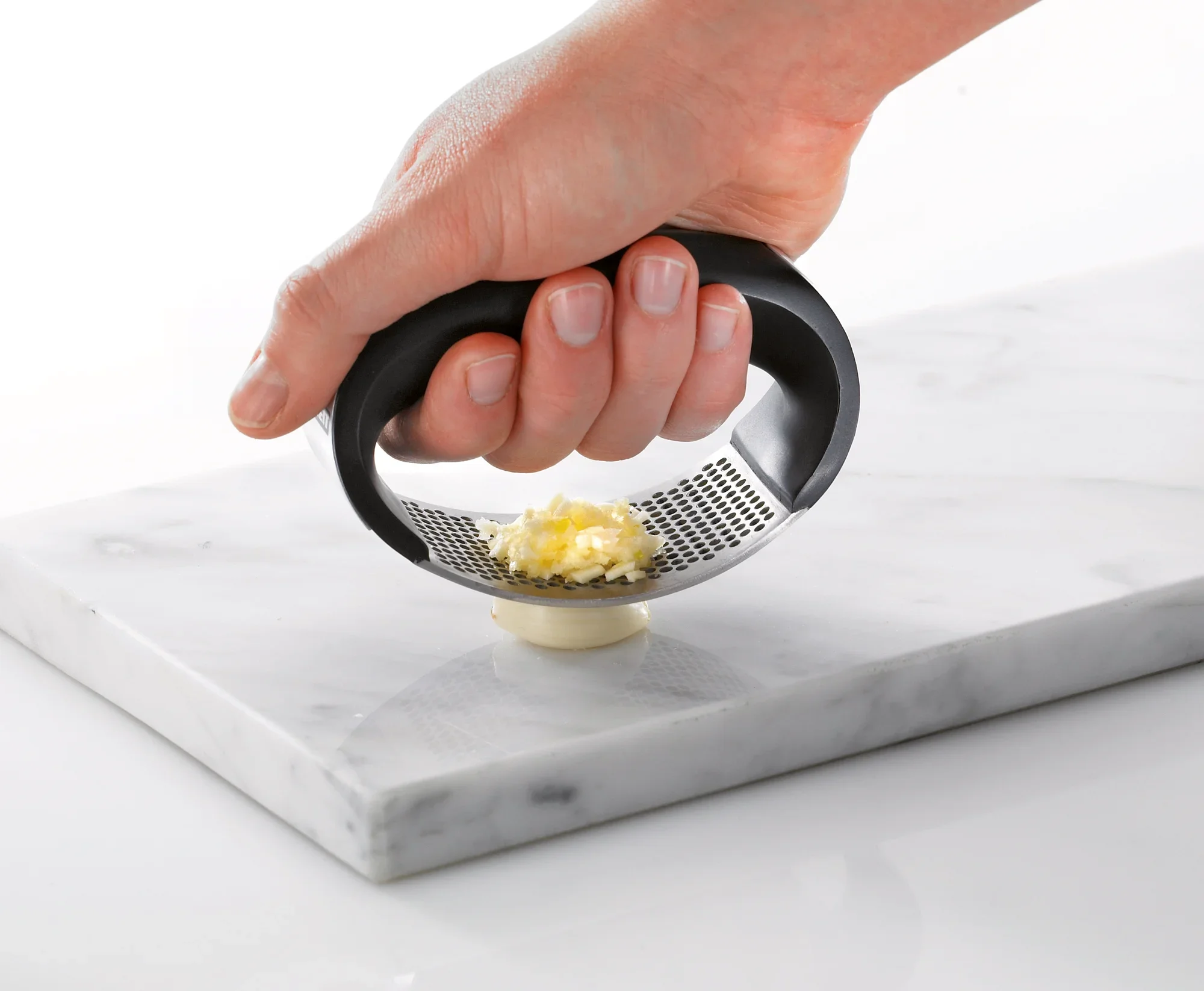 Presse à ail Knobi-King en action : une main appuie le presse-ail ergonomique sur une gousse d'ail pour la broyer sur une planche en marbre. Idéal pour la cuisine.