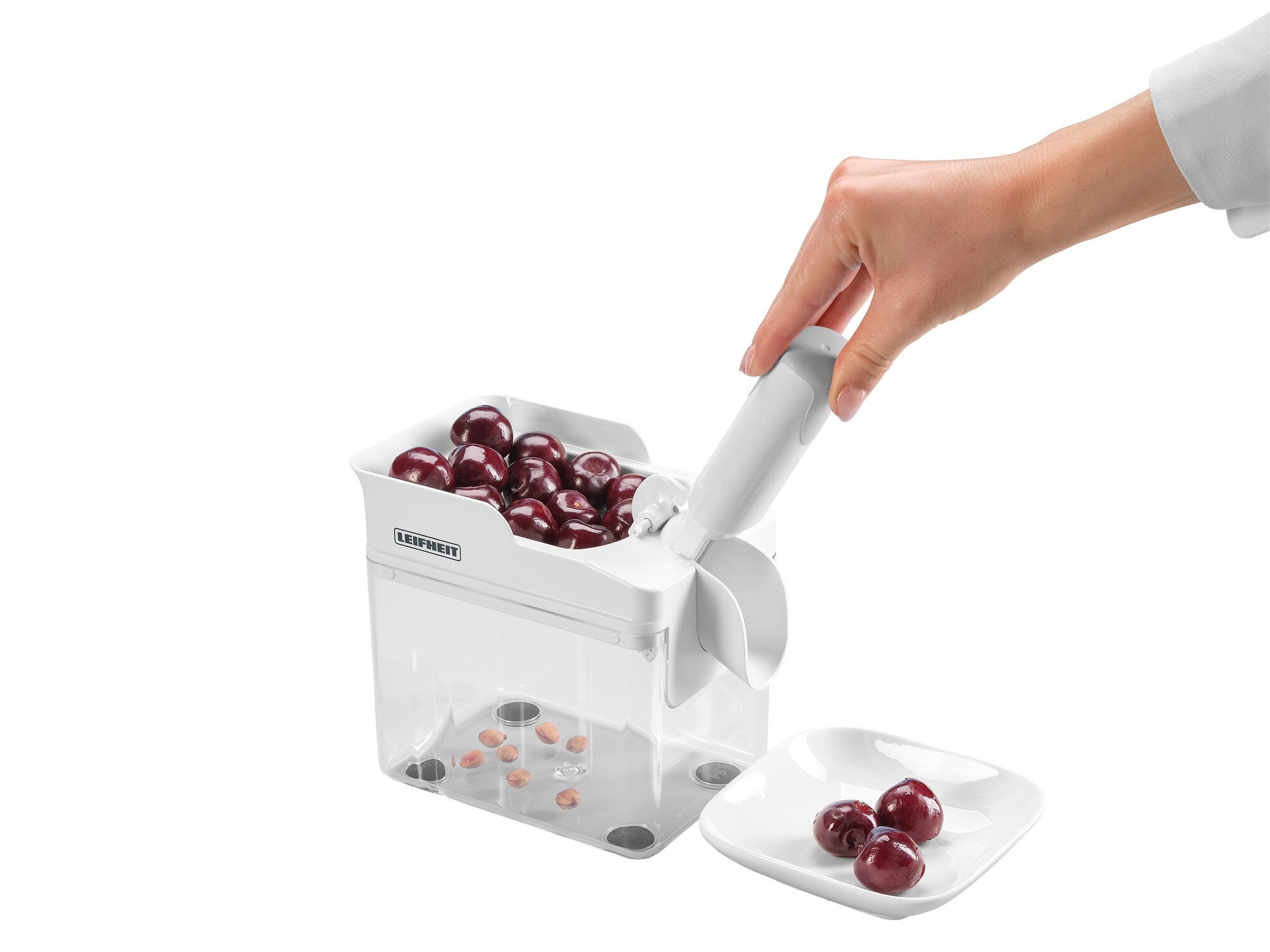 Dénoyauteur de cerises Cherrymat en action : une main actionne le dénoyauteur de cerises blanc, qui dénoyaute les cerises et recueille les noyaux dans un récipient transparent. À côté se trouve une assiette avec des cerises dénoyautées.