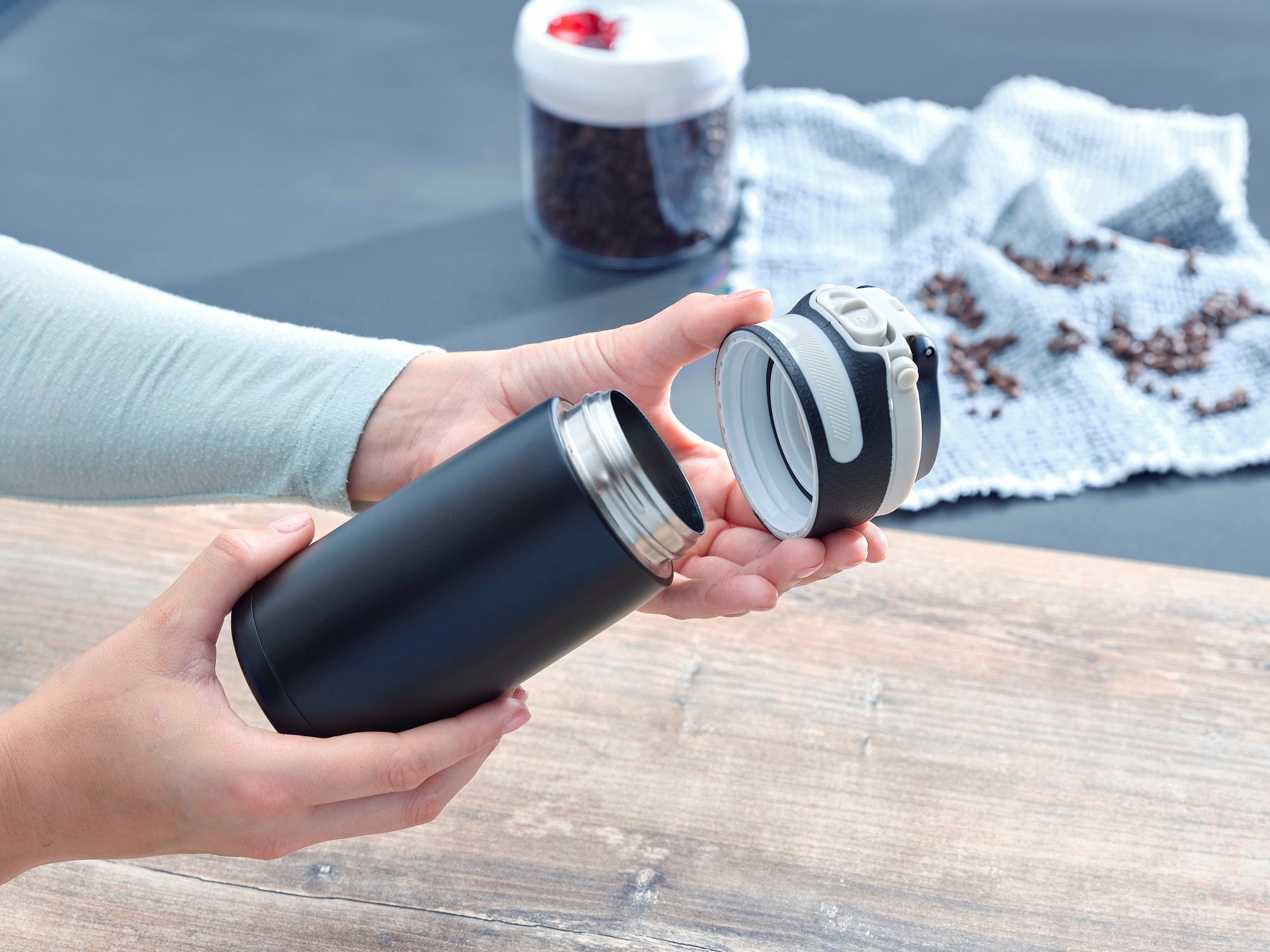 Izolowany kubek Flip 350ml w kolorze czarnym jest otwierany przez jedną osobę. Kubek jest idealny do podróży i utrzymuje napoje gorące lub zimne. W tle widoczne są ziarna kawy.