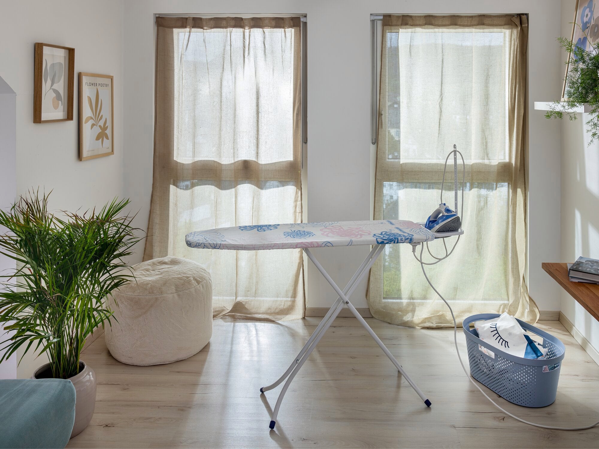 Table à repasser Classic M Basic Plus dans une pièce lumineuse avec deux fenêtres. La table est équipée d'un fer à repasser, à côté se trouve un panier à linge. Des plantes et des tableaux décorent la pièce.