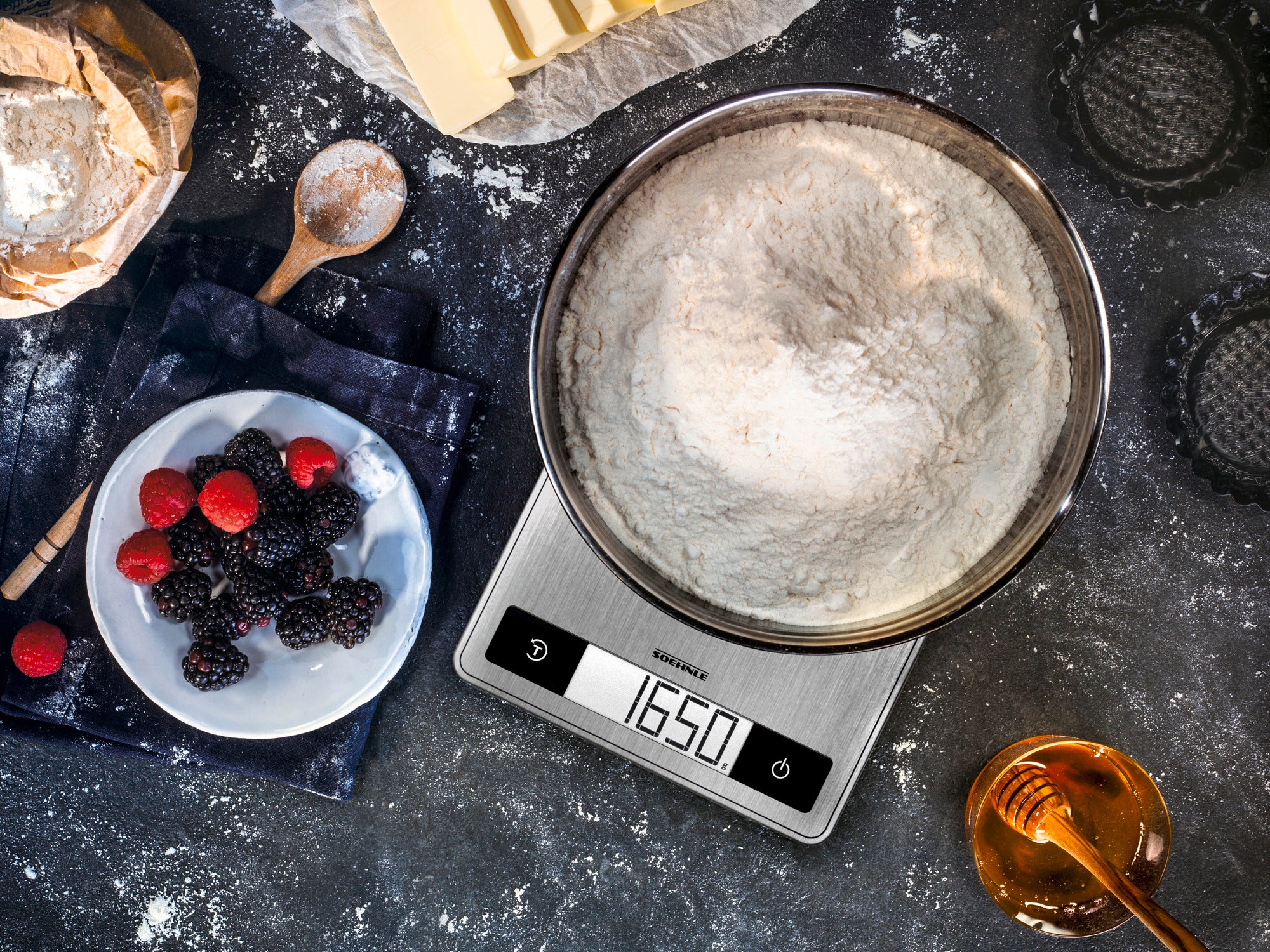 Een digitale keukenweegschaal toont 1650 gram bloem in een metalen kom. Ernaast liggen bessen op een bord, een houten lepel met bloem en een pot honing. Ideaal voor nauwkeurig bakken.
