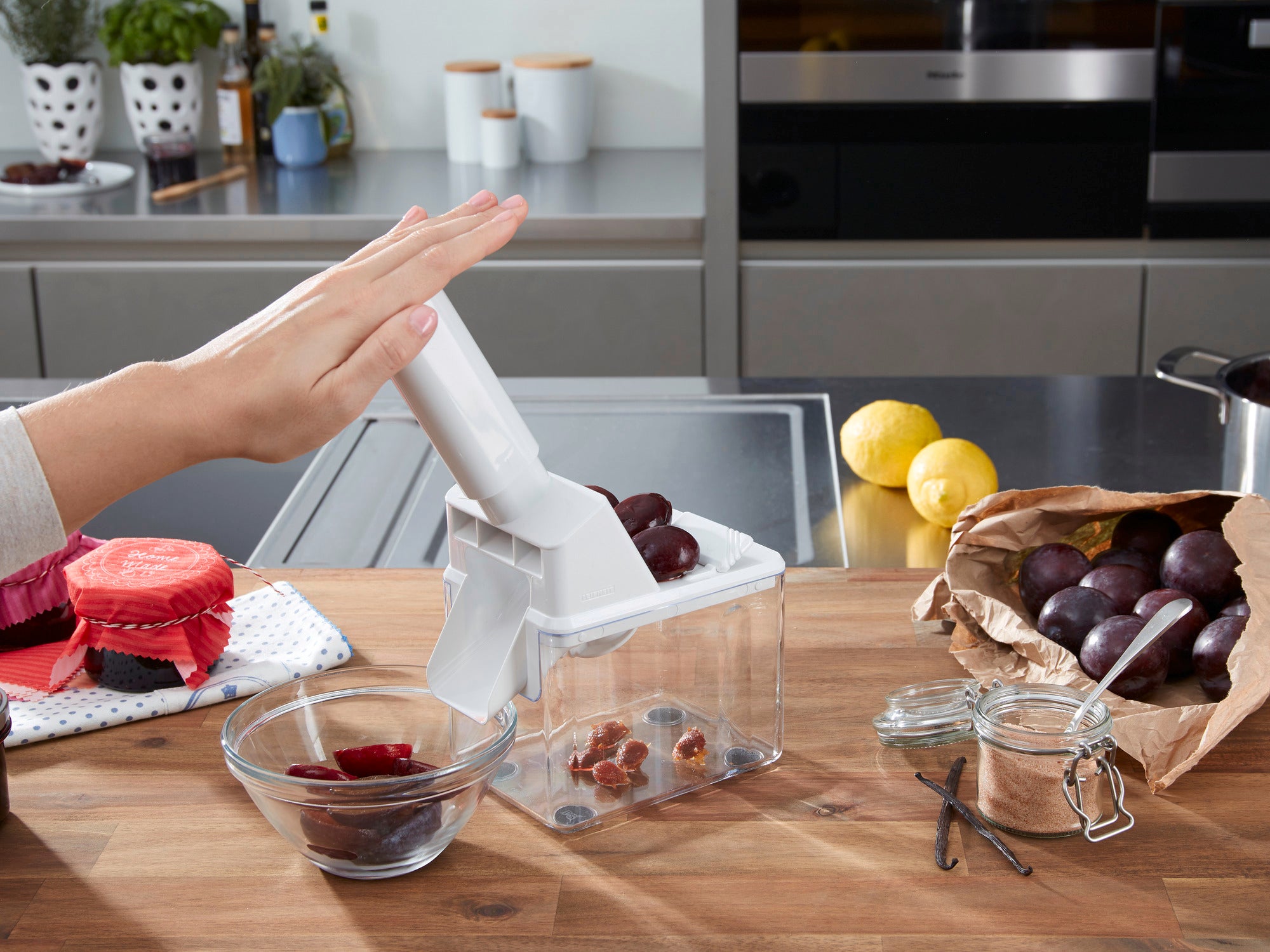 Dénoyauteur de prunes en action sur un plan de travail de cuisine. Une main appuie sur le levier de l'appareil pour dénoyauter les prunes. À côté se trouvent des prunes dénoyautées et un bocal.