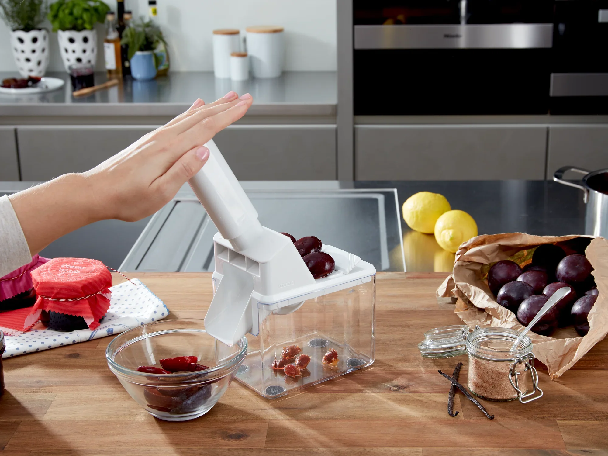 Drylownica do śliwek w użyciu na blacie kuchennym. Ręka naciska dźwignię urządzenia do drylowania śliwek. Obok leżą drylowane śliwki i szklanka.