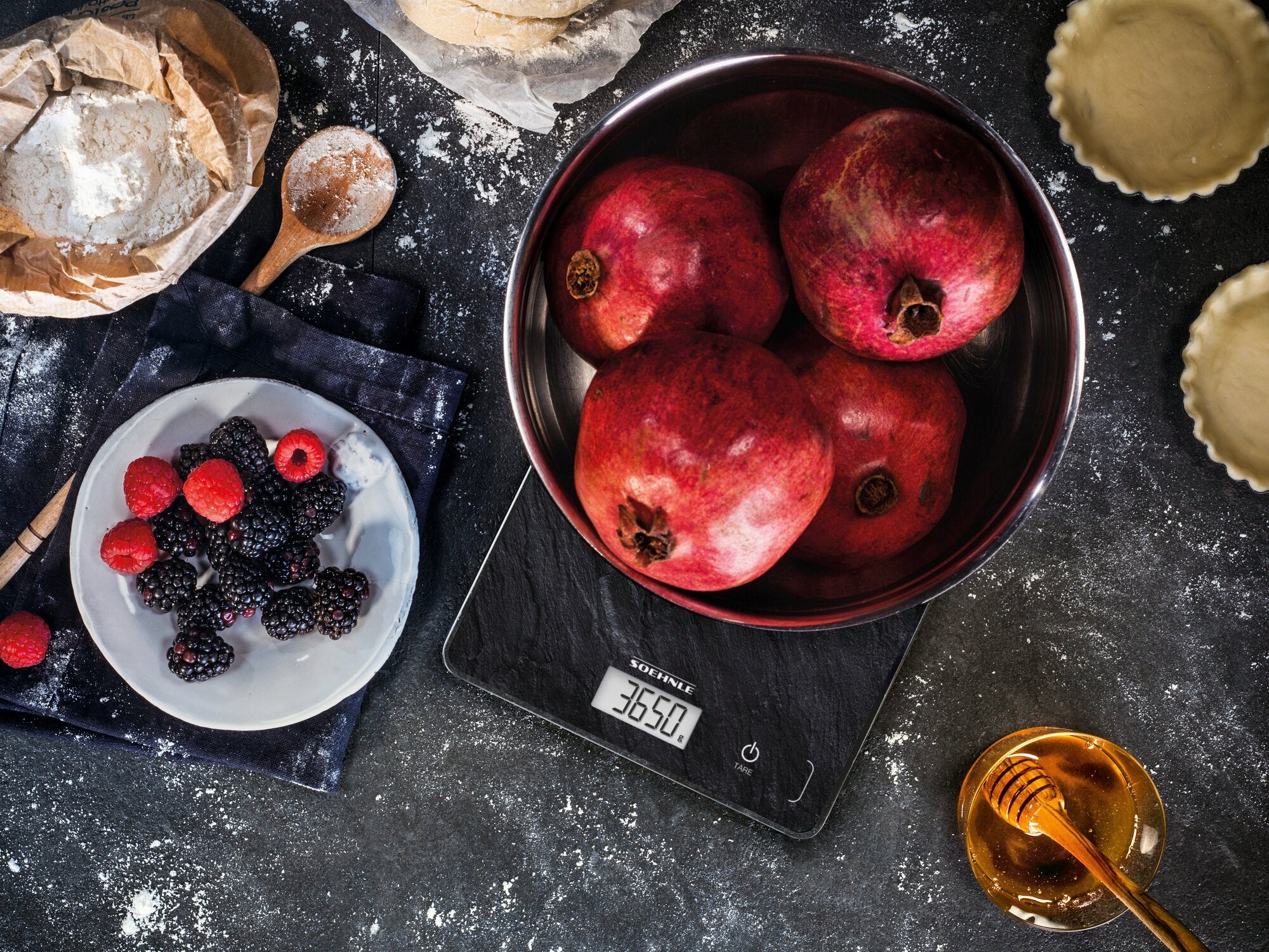 Eine digitale Küchenwaage, Modell Page Compact 300 Slate, zeigt das Gewicht von Granatäpfeln in einer Schüssel an. Daneben liegen Beeren, Mehl und Honig auf einem Tisch.