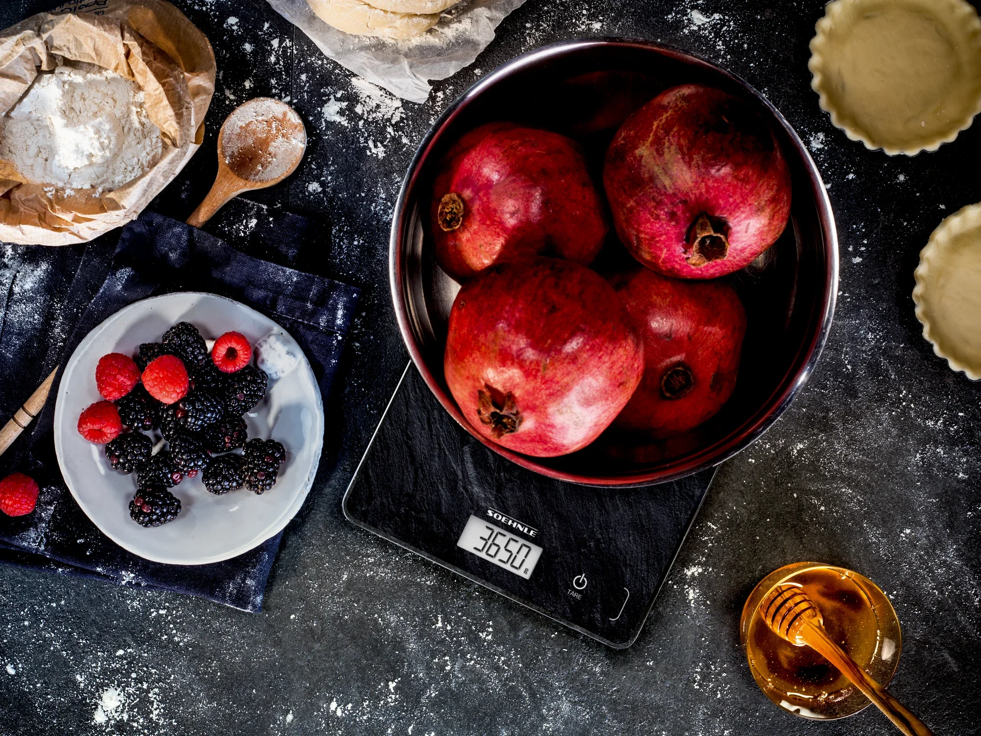 Eine digitale Küchenwaage, Modell Page Compact 300 Slate, zeigt das Gewicht von Granatäpfeln in einer Schüssel an. Daneben liegen Beeren, Mehl und Honig auf einem Tisch.