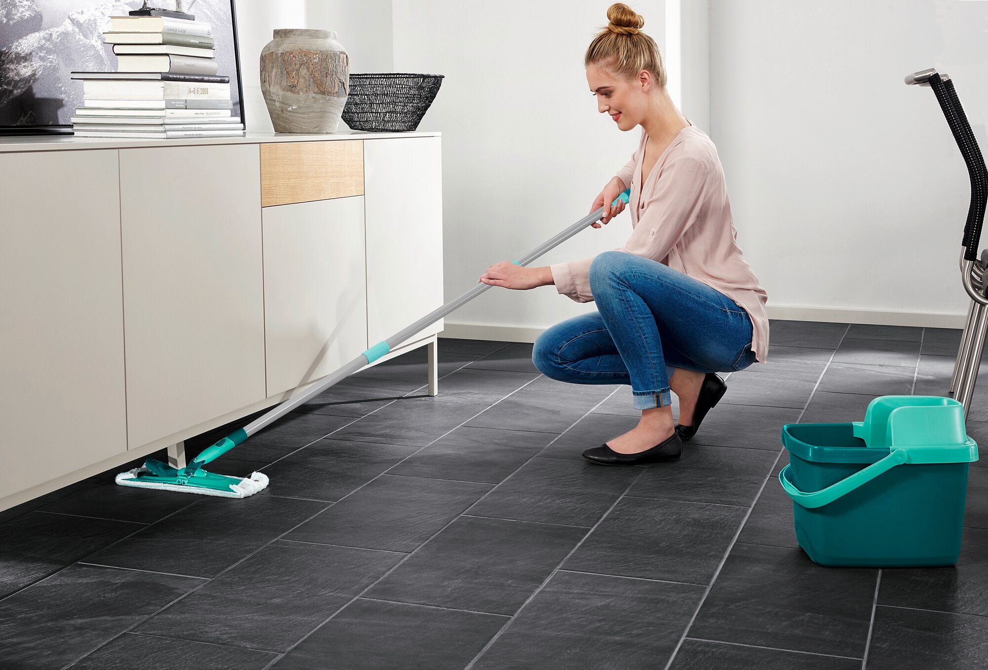 A woman is cleaning the floor with the Combi M floor mop. She is kneeling on a dark tile floor and mopping under a white cabinet. A bucket is standing next to her.