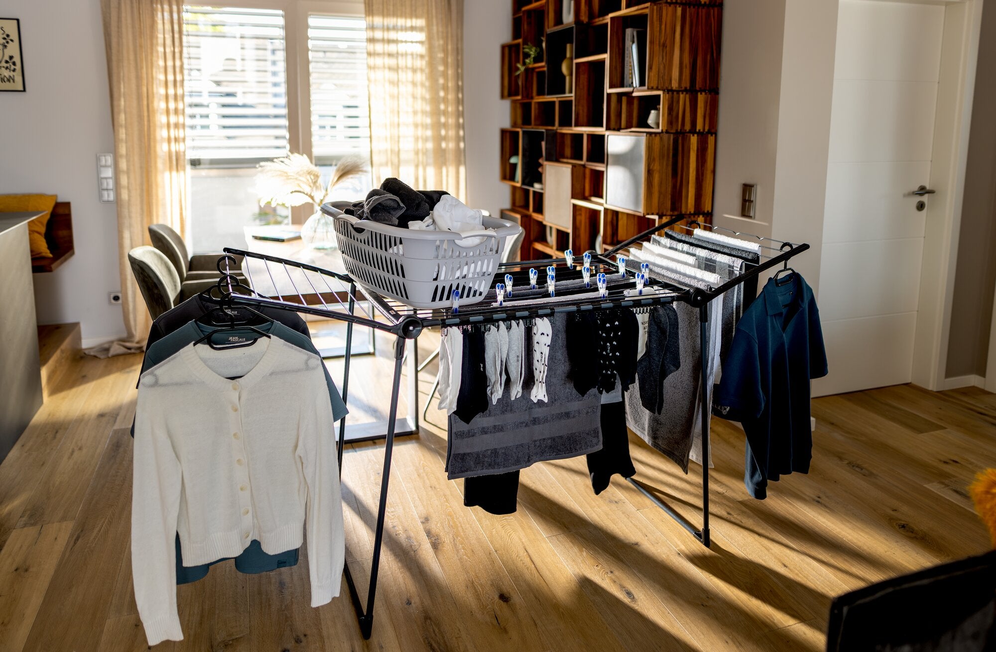 Leifheit Standtrockner Pegasus Ultra Black 23 m in einem hellen Wohnraum, beladen mit Hemden, Handtüchern und dunkler Wäsche; auf den ausklappbaren Flügeln steht ein Wäschekorb.