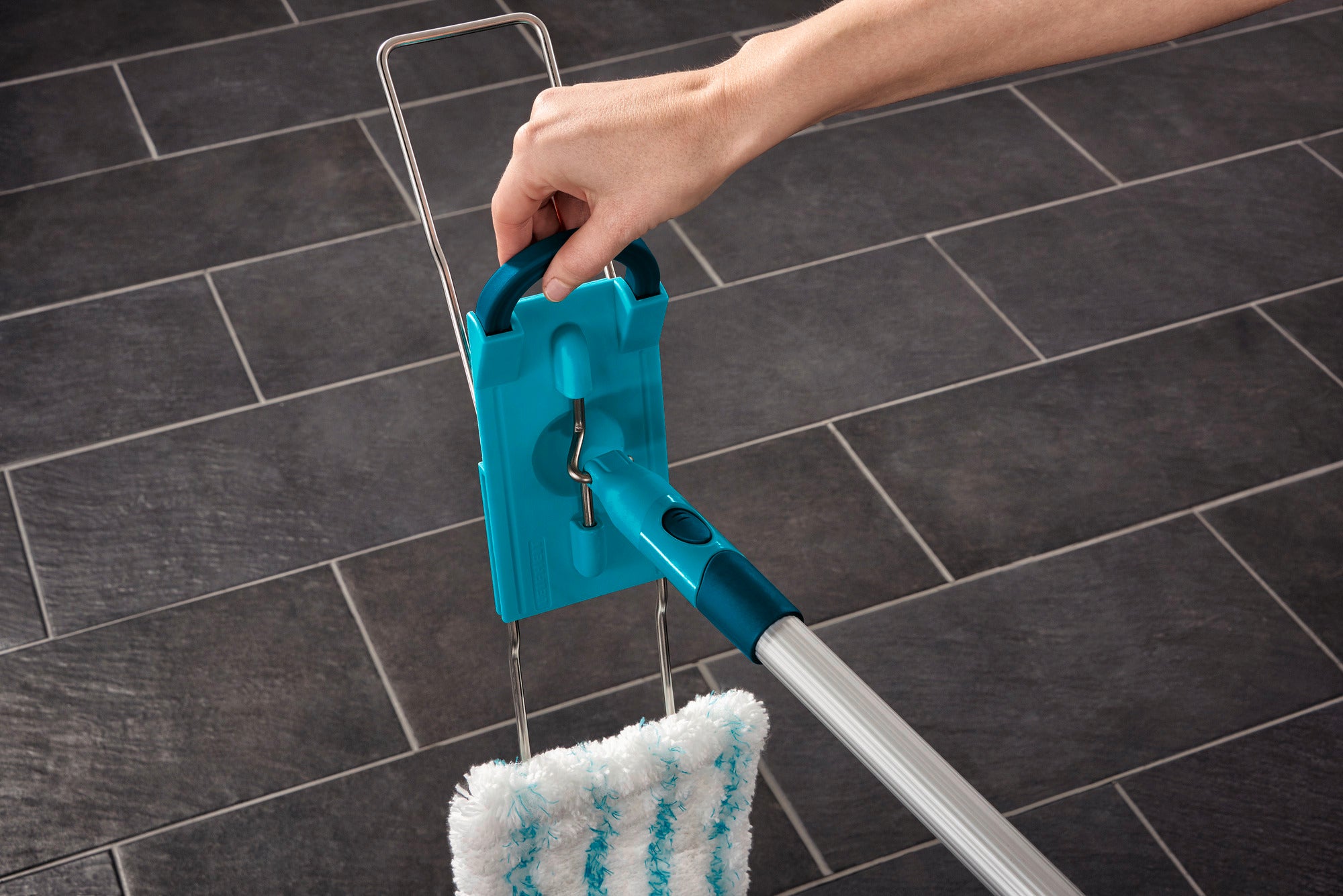 Un balai Profi XL est utilisé sur un sol carrelé foncé. Une main tient le manche du balai, qui est équipé d'un tampon en microfibre bleu et blanc.