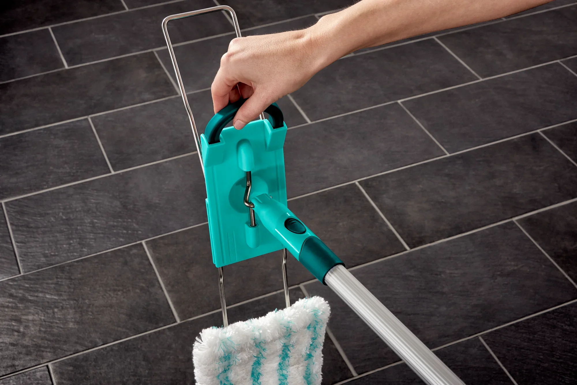 Ein Bodenwischer Profi XL wird auf einem dunklen Fliesenboden verwendet. Eine Hand hält den Griff des Wischers, der mit einem blauen und weißen Mikrofaserpad ausgestattet ist.