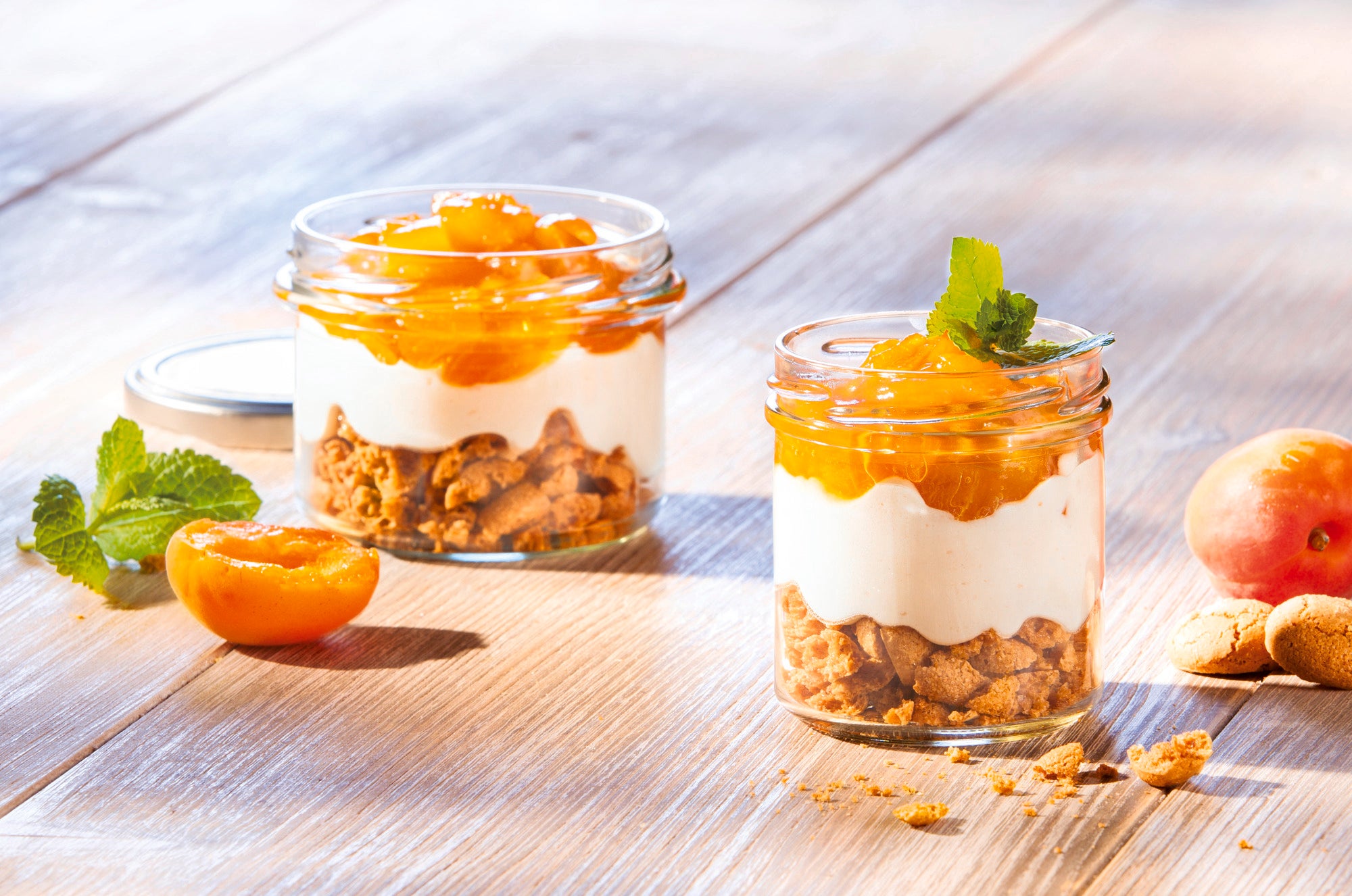 Zwei Sturzgläser, gefüllt mit knusprigem Müsli, Joghurt und frischen Aprikosen, stehen auf einem Holztisch. Ein Glas ist geöffnet, das andere geschlossen. Frische Minzblätter dekorieren die Gläser.