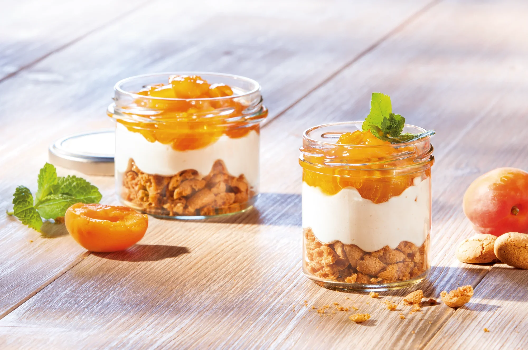 Dwa kubki wypełnione chrupiącym musli, jogurtem i świeżymi morelami stoją na drewnianym stole. Jedna szklanka jest otwarta, druga zamknięta. Szklanki zdobią liście świeżej mięty.