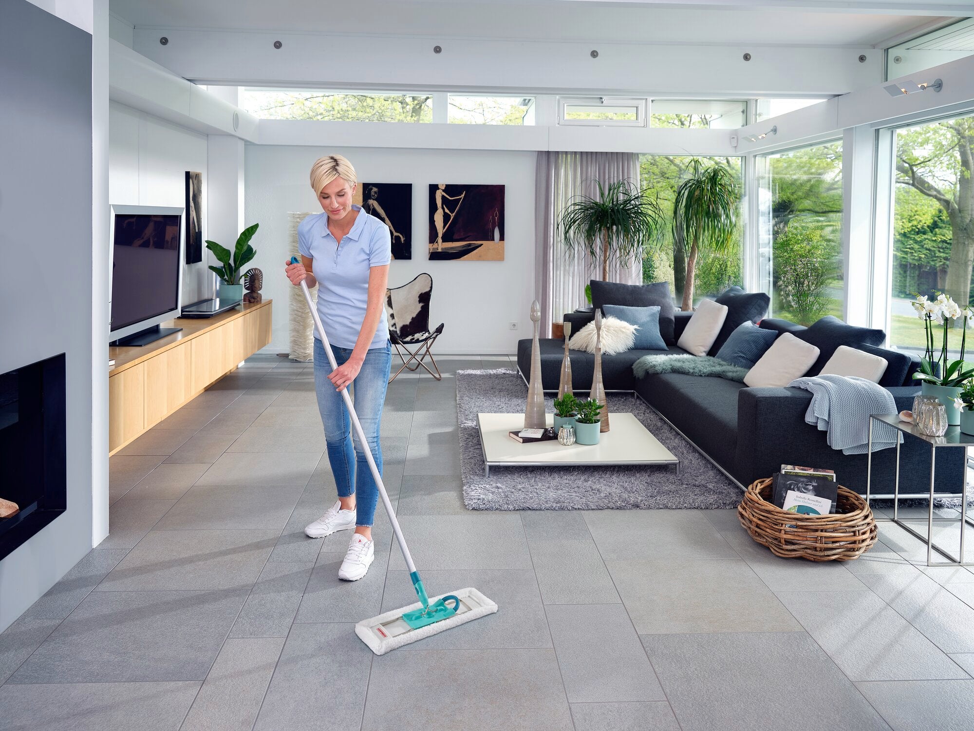 Een vrouw dweilt de vloer van een moderne woonkamer met de SUPERDUSTER Floor XL. De kamer is licht en stijlvol, met grote ramen en gezellige meubels.