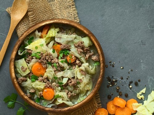 Eine Holzschale mit Wirsing-Eintopf, garniert mit Hackfleisch, Karottenscheiben und Petersilie, auf einem grauen Untergrund mit Holzloeffel.