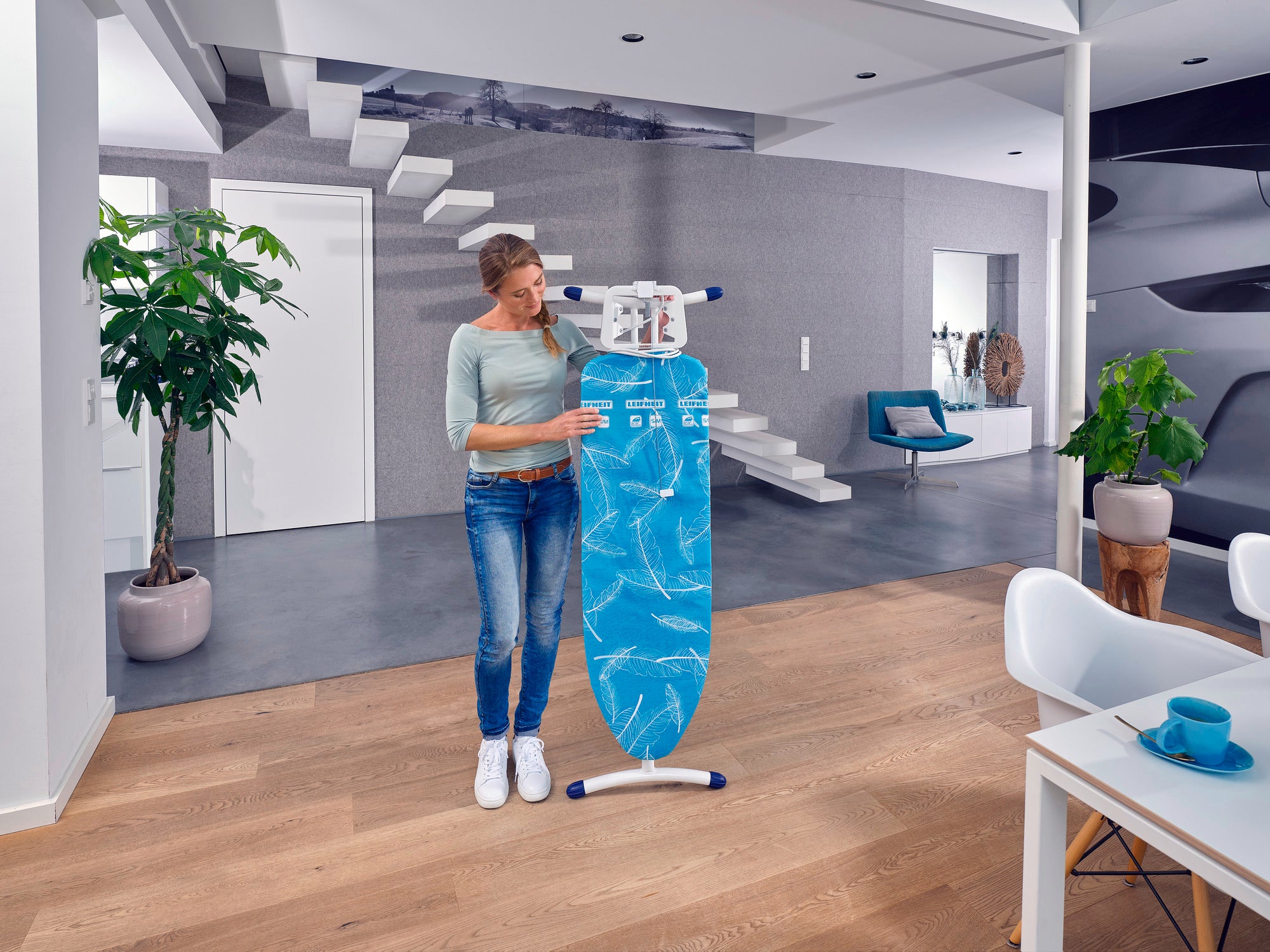 Eine Frau steht in einem modernen Wohnzimmer und hält einen Bügeltisch Air Board M Solid Plus. Der Raum ist stilvoll eingerichtet mit Pflanzen und modernen Möbeln.
