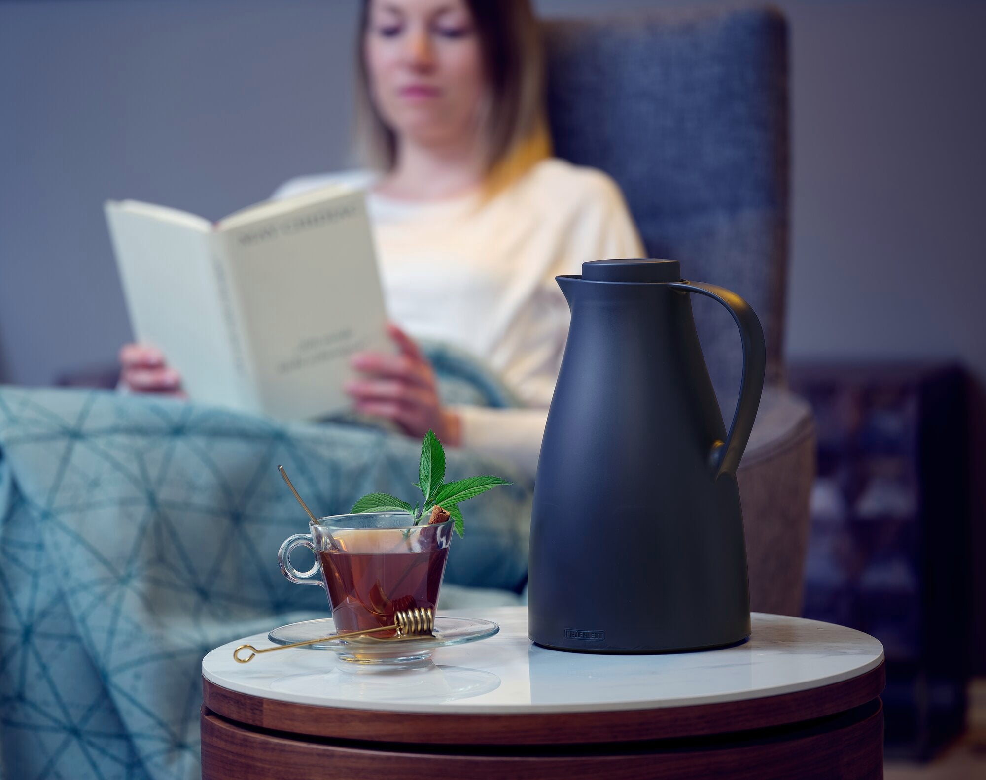 Isolierkanne Harmonic auf einem Tisch neben einer Tasse Tee. Im Hintergrund sitzt eine Person und liest ein Buch. Die Szene vermittelt Ruhe und Entspannung.