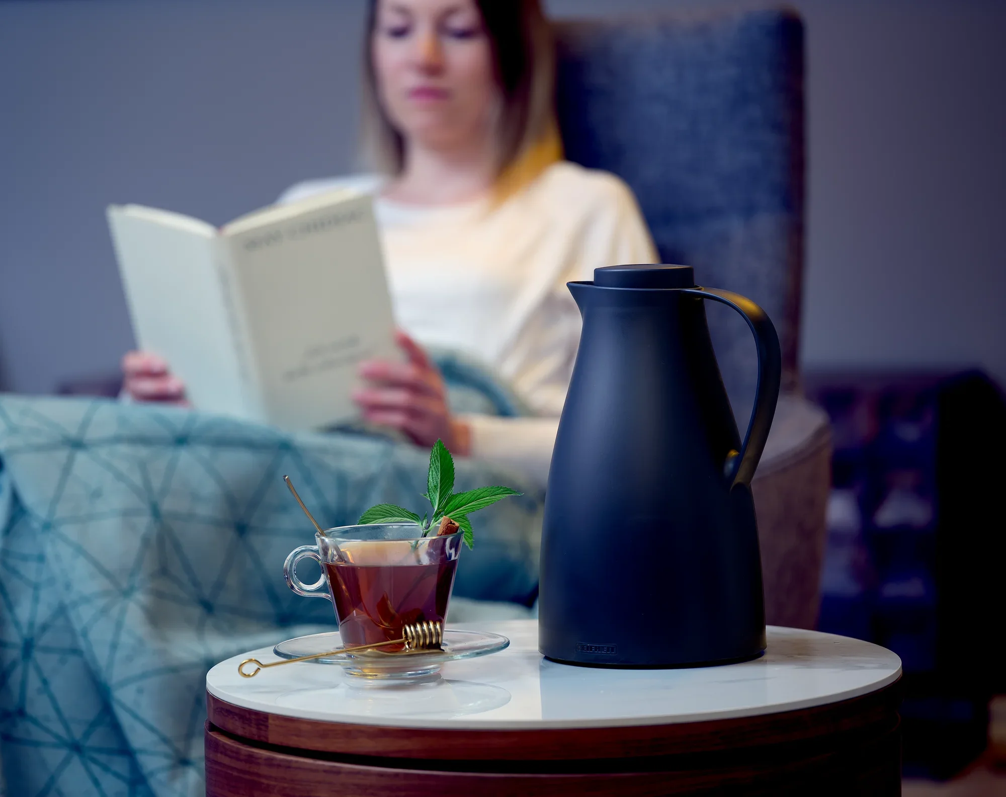 Harmonische isoleerkan op een tafel naast een kopje thee. Een persoon zit op de achtergrond een boek te lezen. Het tafereel straalt rust en ontspanning uit.
