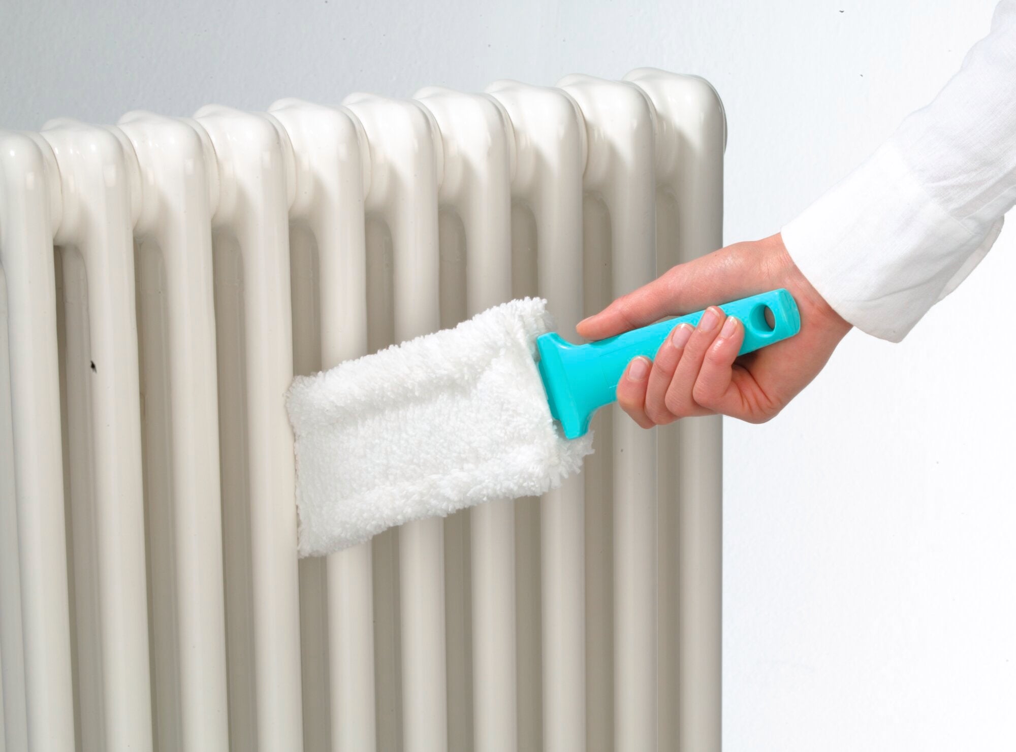 Eine Hand benutzt den Heizkörperreiniger Duster Flat, um Staub zwischen den Lamellen eines weißen Heizkörpers zu entfernen. Der Reiniger hat einen blauen Griff und ein weißes Reinigungspad.