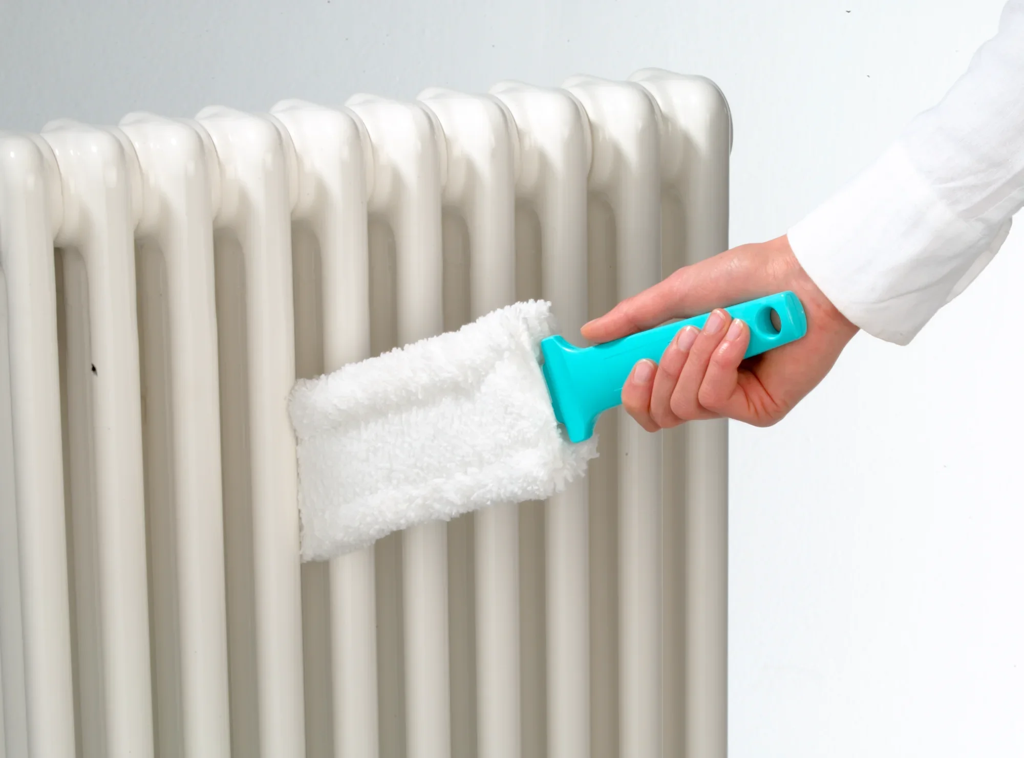 Eén hand gebruikt de Duster Flat radiatorreiniger om stof te verwijderen tussen de lamellen van een witte radiator. De reiniger heeft een blauw handvat en een witte reinigingspad.
