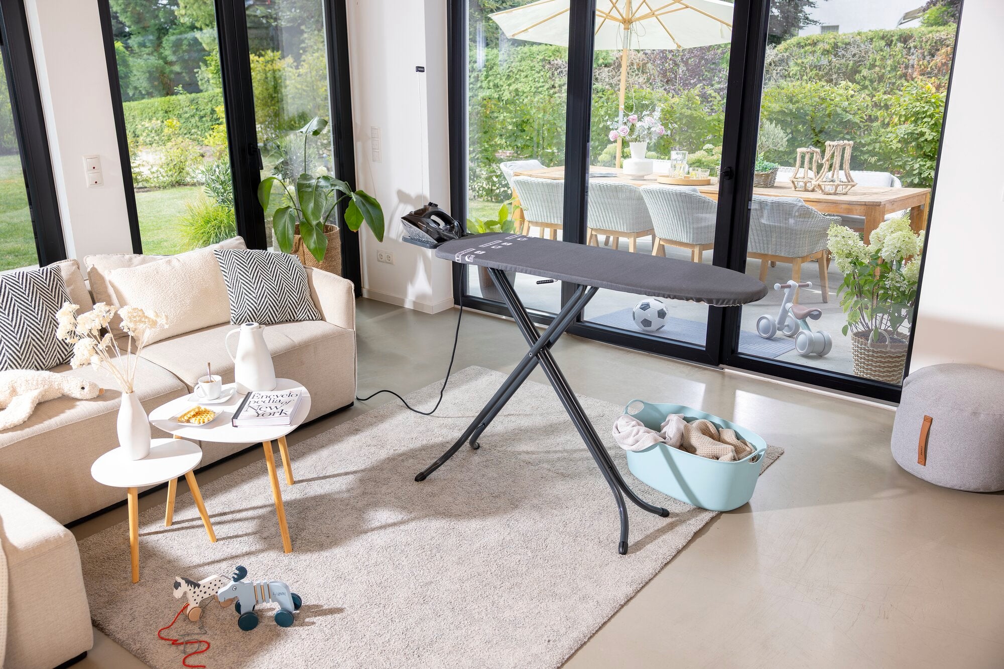 Bügelbrett Leifheit Air Board M Black in einem modernen Wohnzimmer. Das Brett steht auf einem hellen Teppich, umgeben von einem Sofa, Tisch und Spielzeug. Im Hintergrund ist ein großer Garten sichtbar.