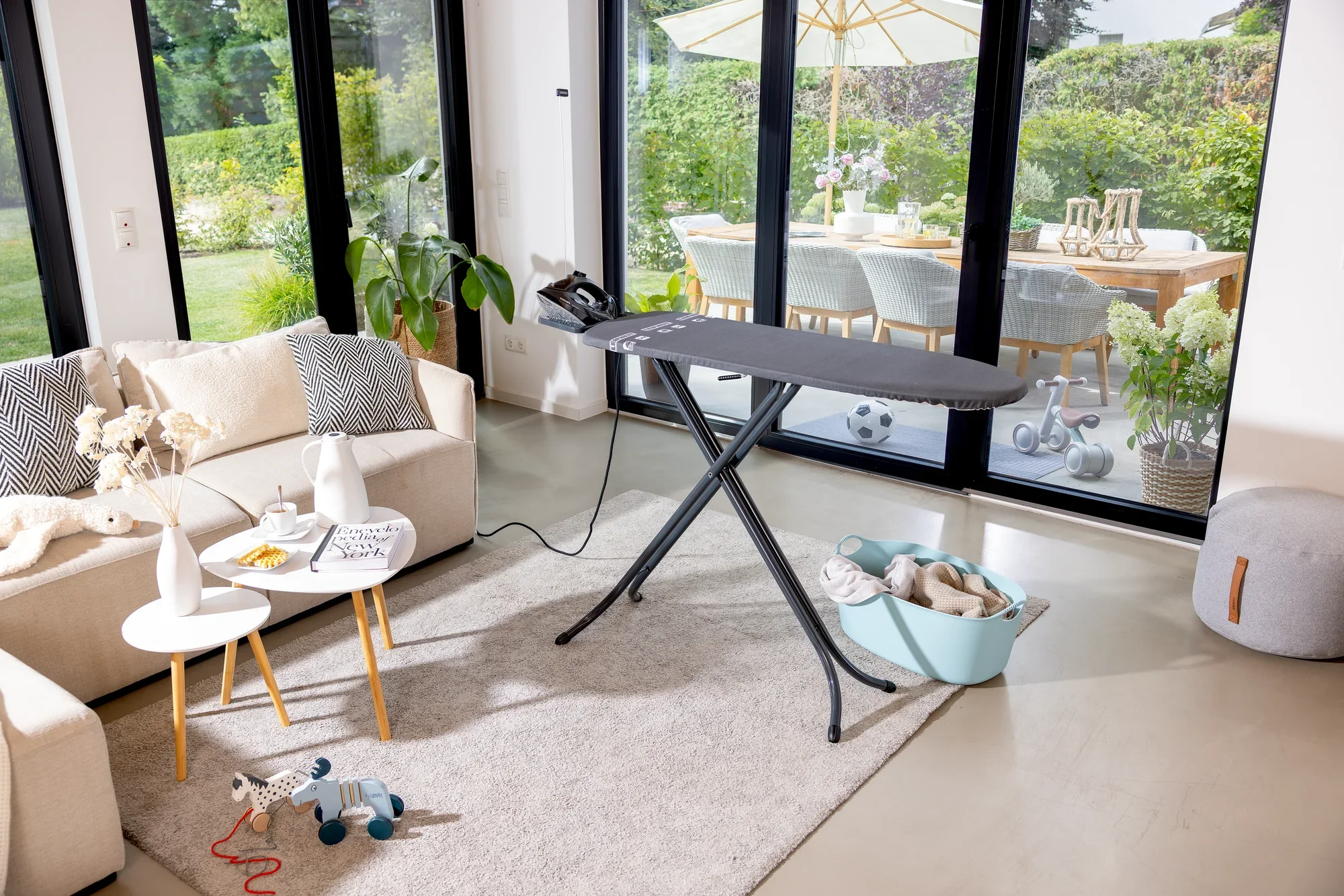 Bügelbrett Leifheit Air Board M Black in einem modernen Wohnzimmer. Das Brett steht auf einem hellen Teppich, umgeben von einem Sofa, Tisch und Spielzeug. Im Hintergrund ist ein großer Garten sichtbar.