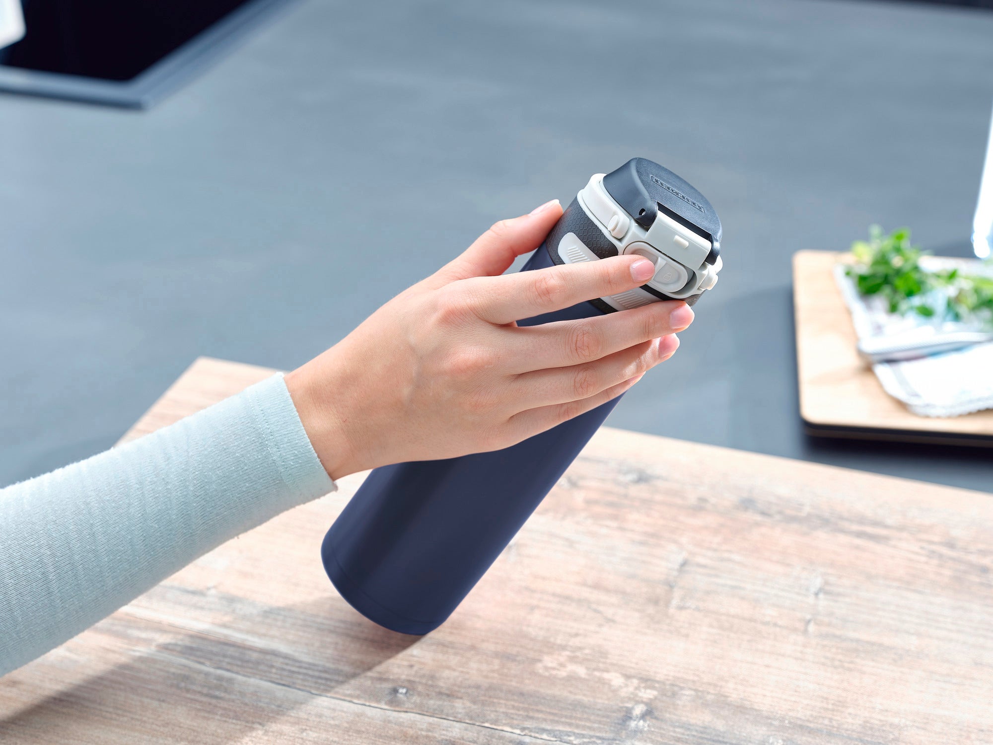 Une main tient un mug isotherme Flip bleu foncé d'une contenance de 600 ml. Le mug est posé sur une table en bois, des ustensiles de cuisine sont visibles à l'arrière-plan.