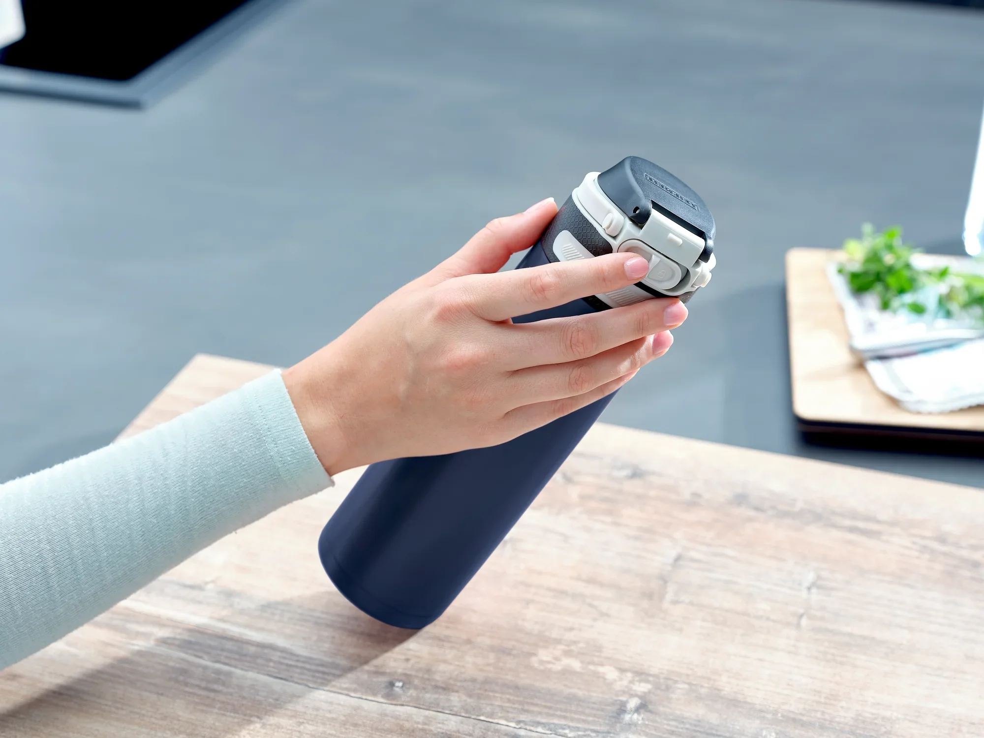 Una mano sostiene una taza térmica Flip azul oscuro con una capacidad de 600 ml. La taza está sobre una mesa de madera y al fondo se ven utensilios de cocina.
