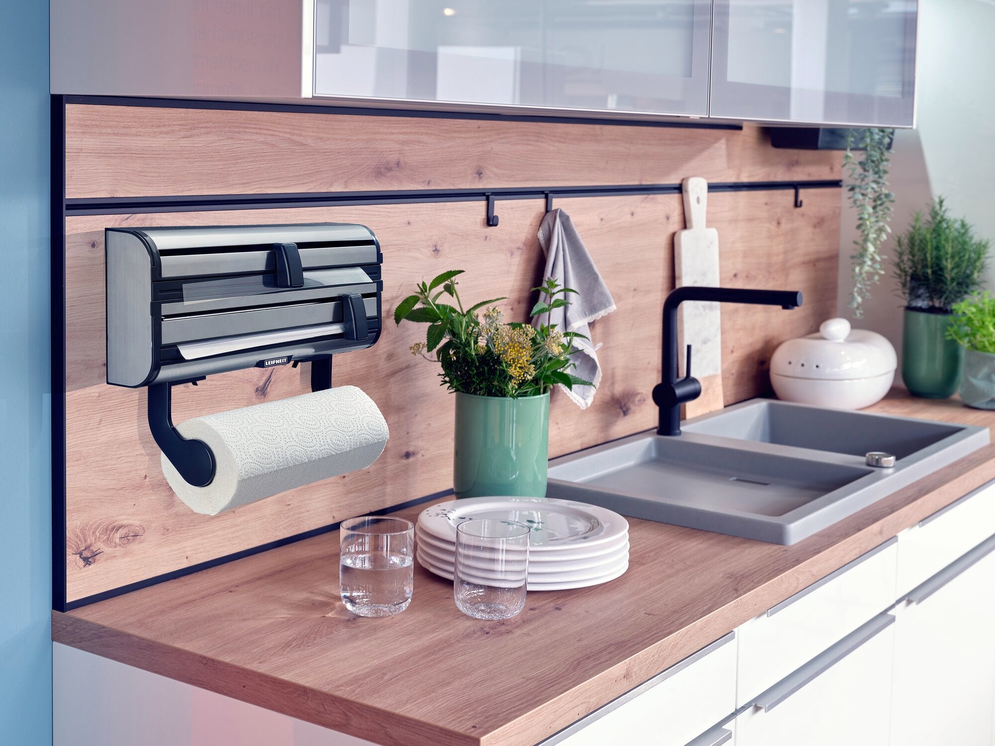 Parat Royal wall-mounted roll holder installed in a modern kitchen. The holder is attached to a wooden back panel and holds a kitchen roll. Next to it are plants and crockery.
