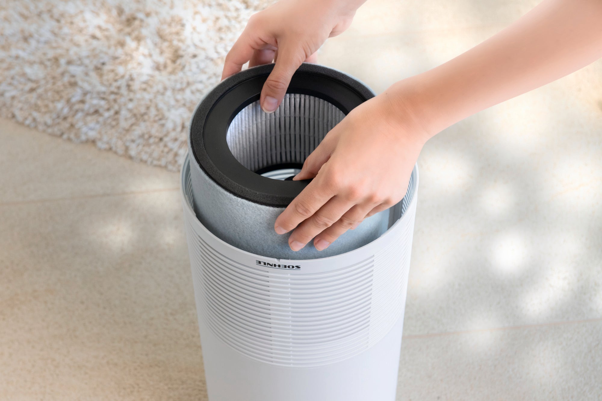 Een persoon verwisselt het filter van een Airfresh Clean 400 luchtreiniger. De luchtreiniger staat op een lichtgekleurde vloer en op de achtergrond is een tapijt te zien.