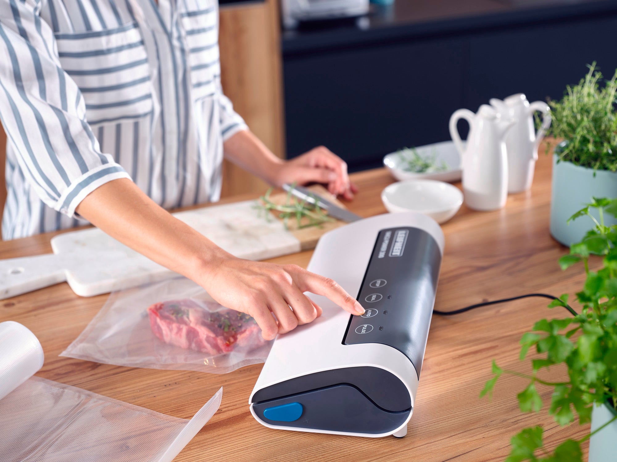 Eine Person verwendet das Vakuumiergerät Vacu Power 300, um Lebensmittel zu versiegeln. Das Gerät liegt auf einer Holzarbeitsplatte, umgeben von frischen Kräutern und Geschirr.