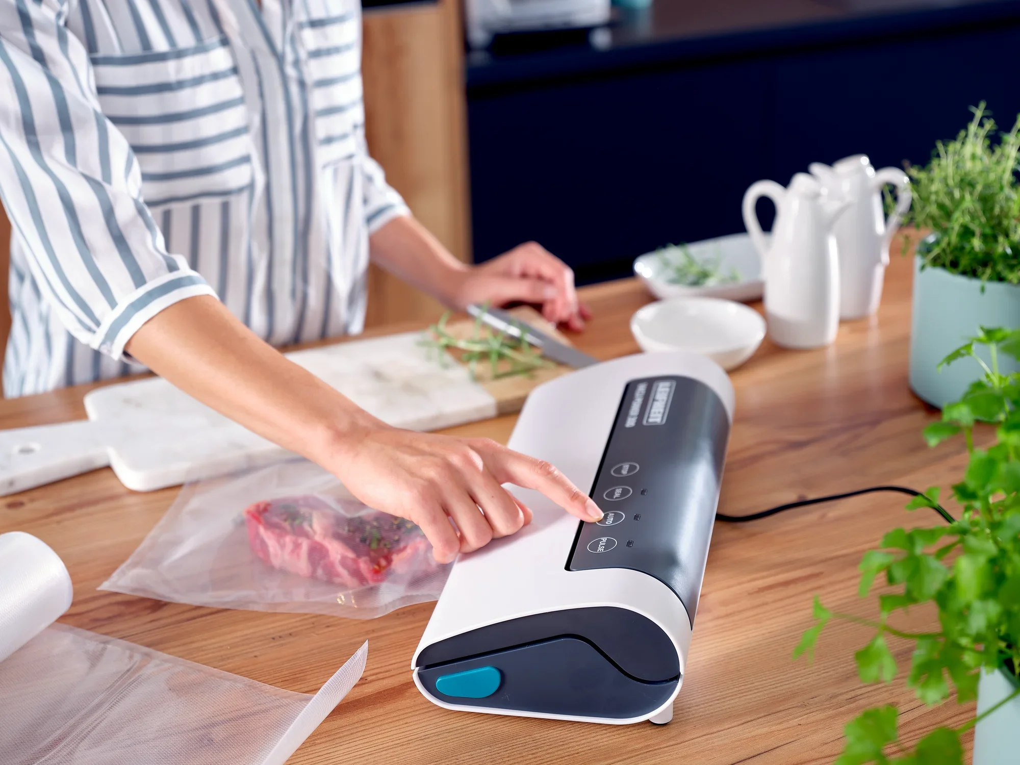 Une personne utilise l'appareil de mise sous vide Vacu Power 300 pour sceller des aliments. L'appareil est posé sur un plan de travail en bois, entouré d'herbes fraîches et de vaisselle.