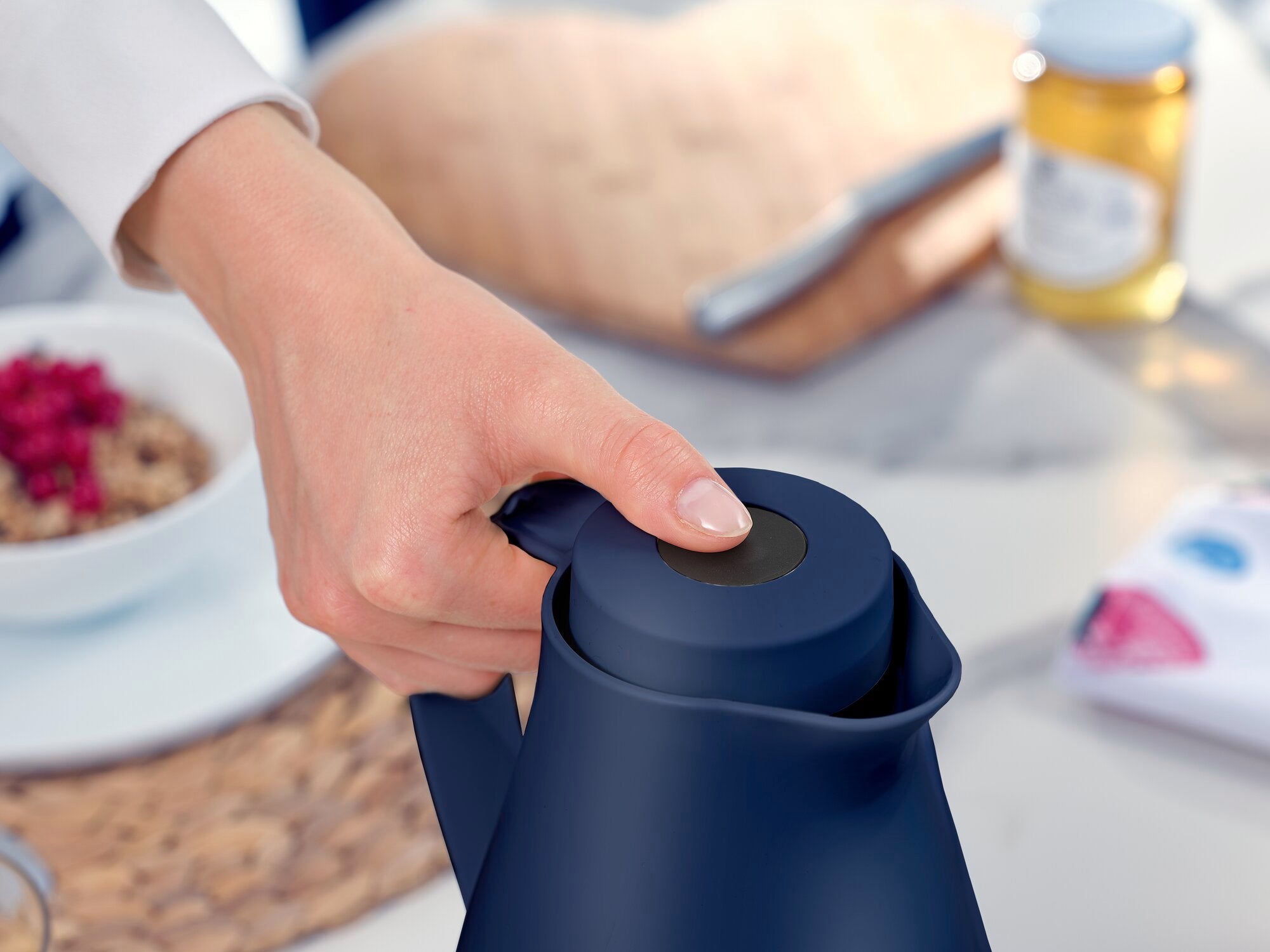 Thermos Harmonic en cours d'utilisation. Une main tient le couvercle de la verseuse bleu foncé, tandis qu'à l'arrière-plan, on aperçoit une table de petit-déjeuner avec des céréales et du miel.