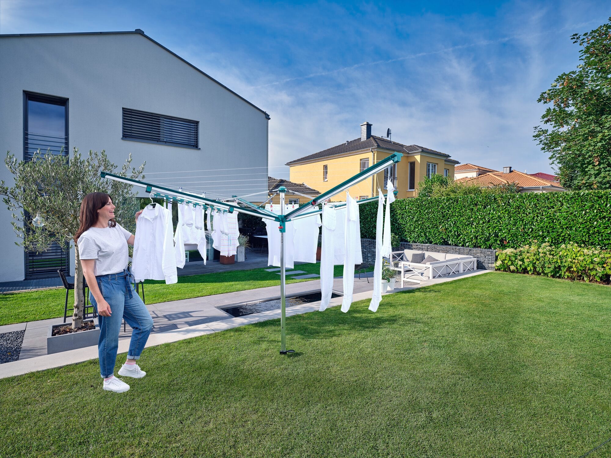 Eine Frau steht in einem Garten neben einem Wäscheschirm, auf dem weiße Kleidung hängt. Der Easy Clip Kleinteilehalter von Linomatic wird verwendet, um die Wäsche effizient zu trocknen.
