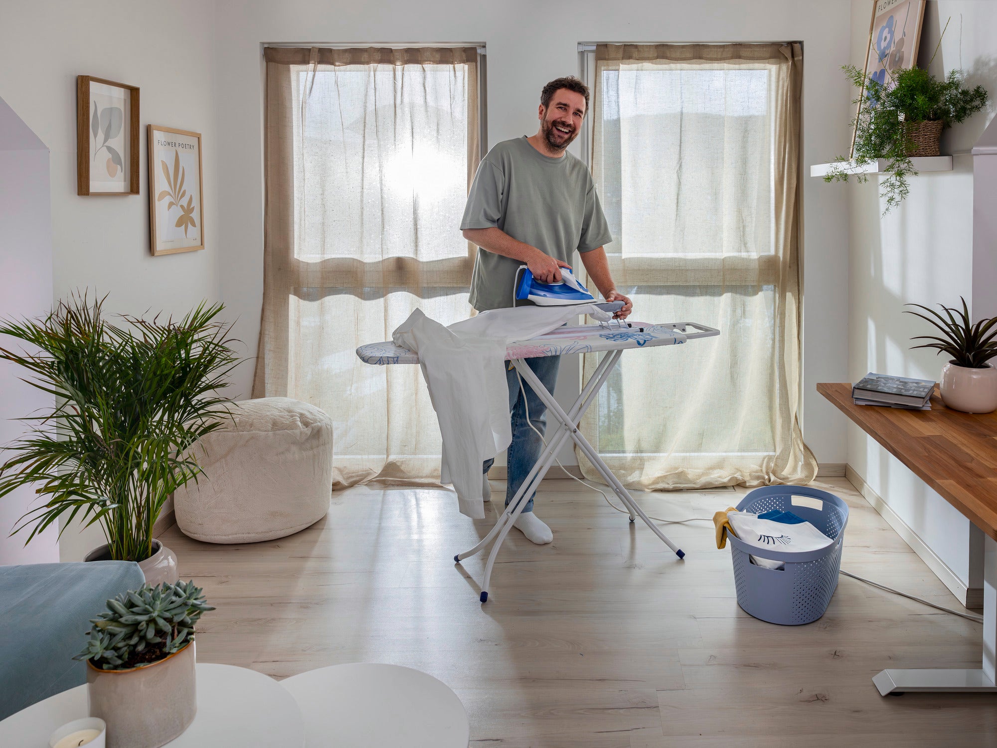 Un hombre plancha una camisa en una tabla de planchar en una habitación luminosa y amueblada de forma moderna. Al fondo se ven grandes ventanas con cortinas y plantas.